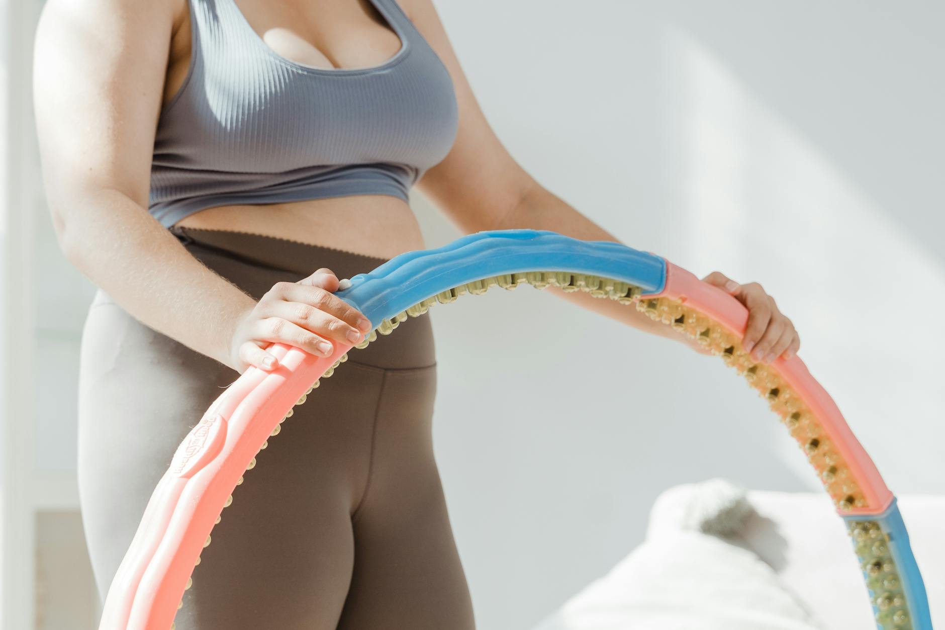 A woman in a gray sports bra and beige leggings holding a pink and blue hula hoop. She is standing in a room with a white wal...