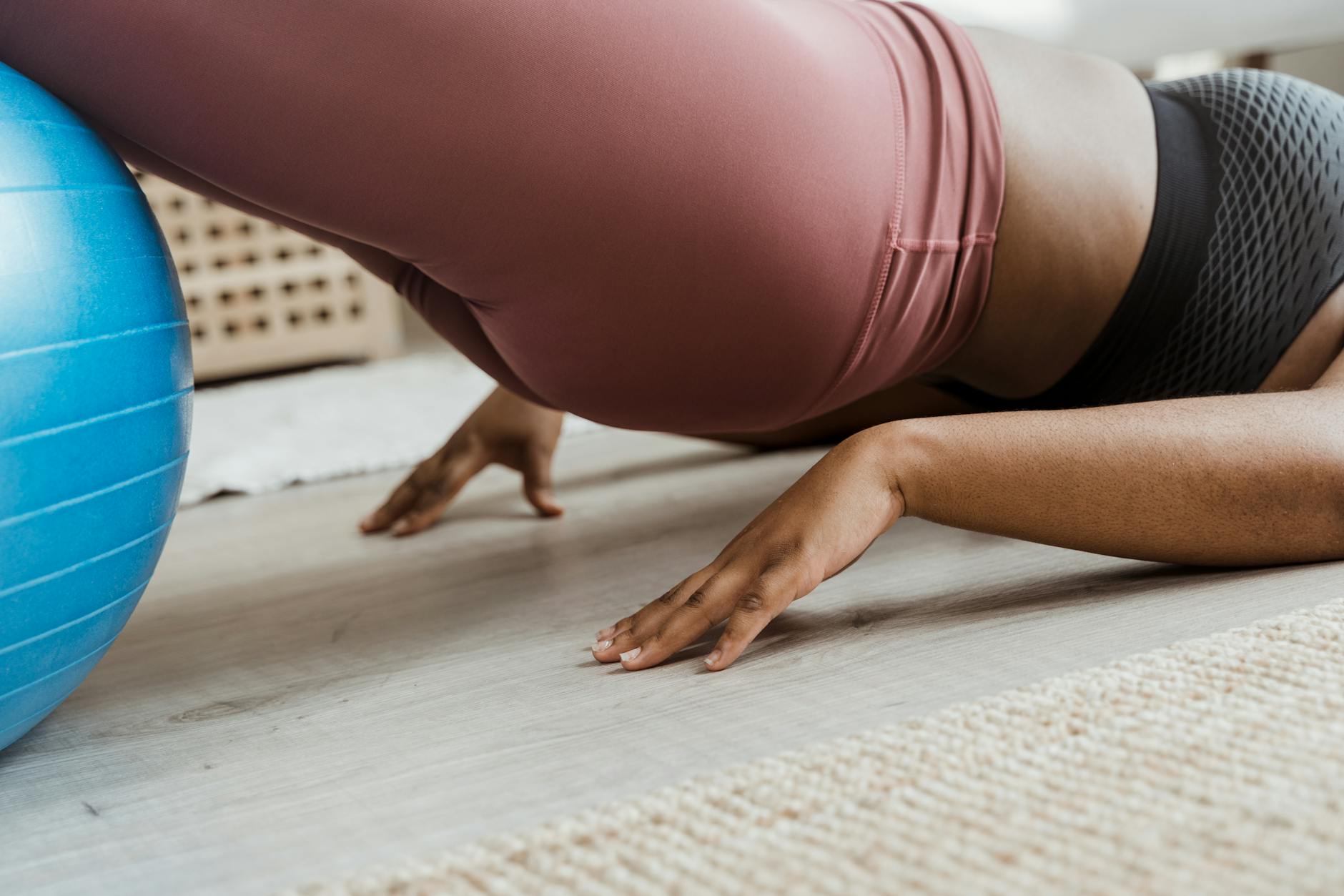 A woman performing a plank exercise on a blue exercise ball. She is wearing a pink sports bra and black leggings. Her arms ar...
