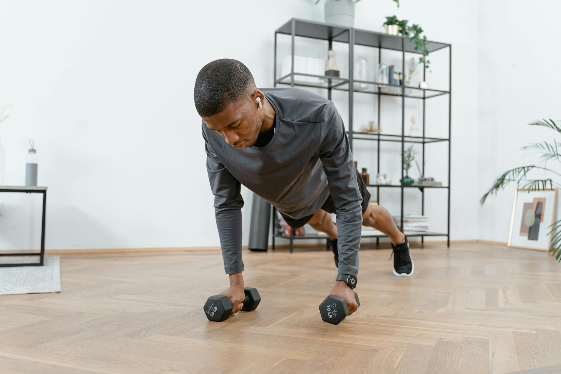 A young man in a living room, performing a push-up exercise with dumbbells. He is wearing a grey long-sleeved shirt and black...