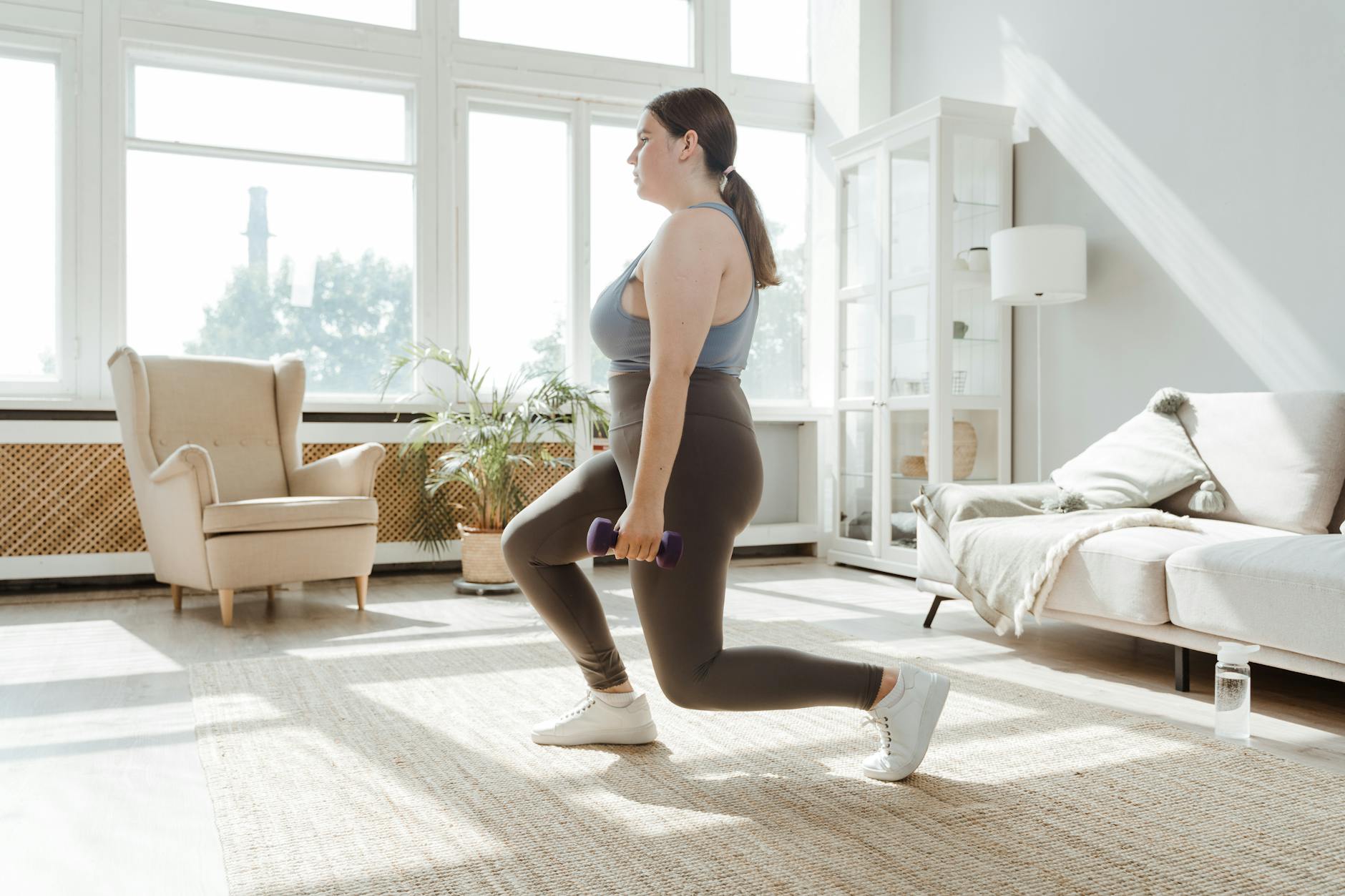 A young woman in a living room, performing a lunge exercise with a purple dumbbell. She is wearing a gray tank top, grey legg...