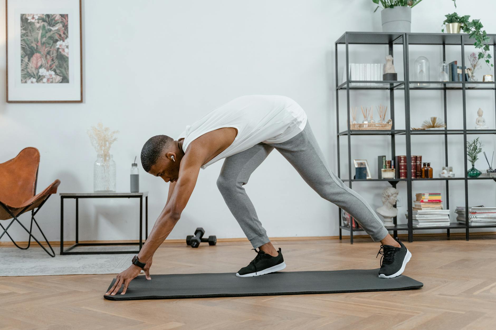 A man in a white tank top and grey sweatpants performing a plank exercise on a black yoga mat in a living room. He is in a si...
