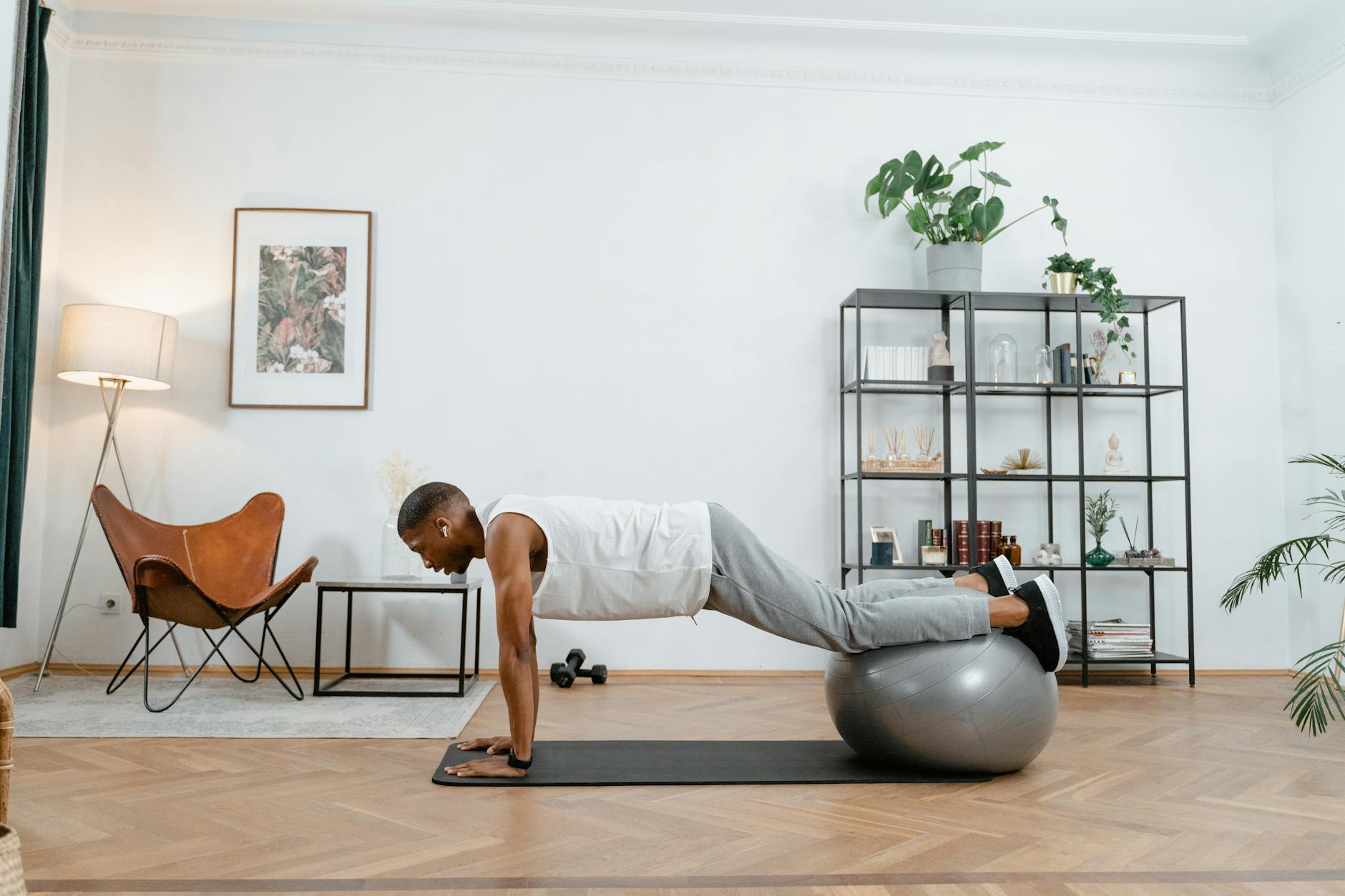 A young man performing a plank exercise on an exercise ball in a living room. He is wearing a white tank top and grey sweatpa...