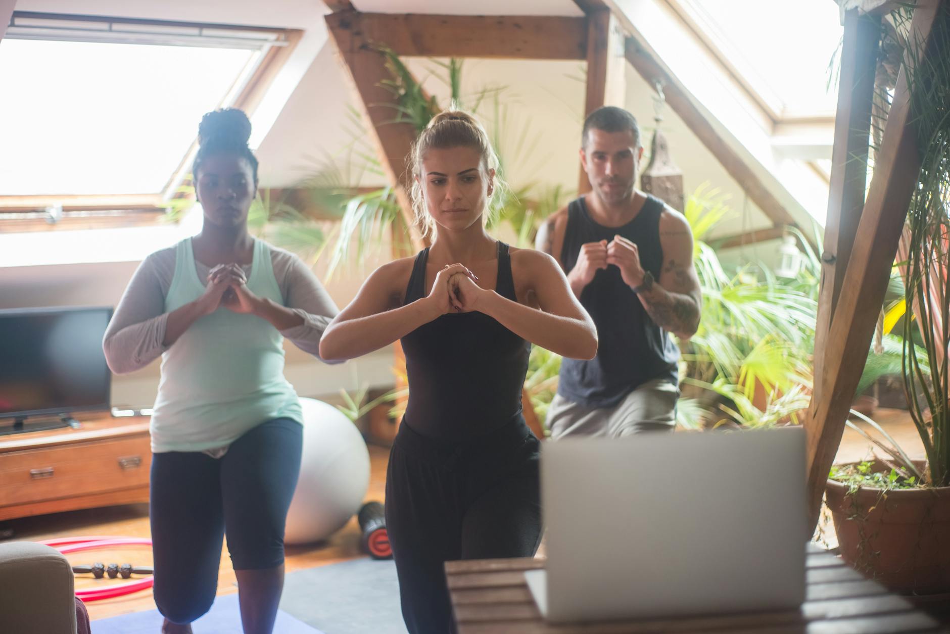 Three people in a yoga class in a room with a high ceiling and large windows. They are standing in a line with their hands cl...