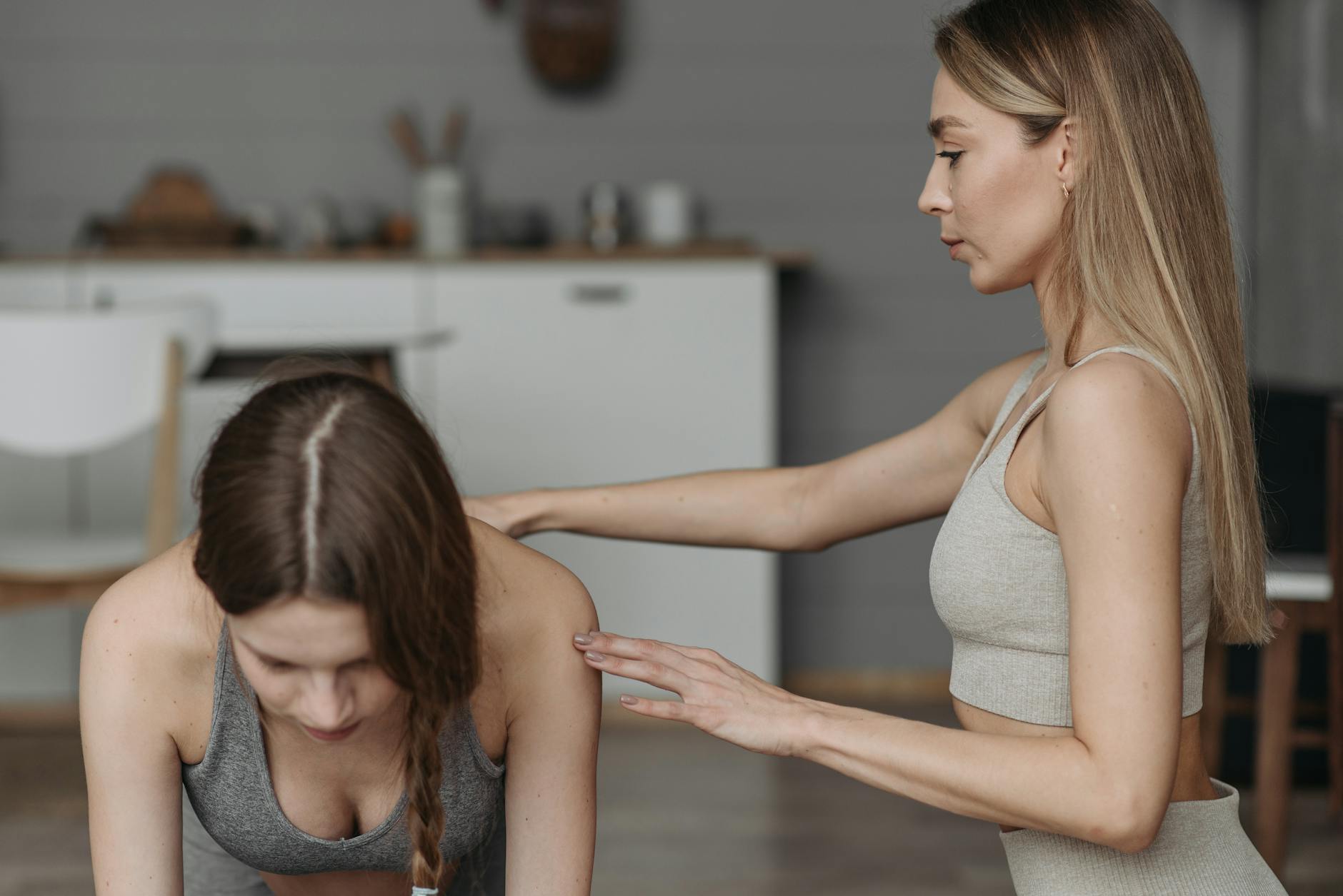 Two young women in a kitchen. The woman on the left is kneeling on the floor with her back to the camera, wearing a gray spor...