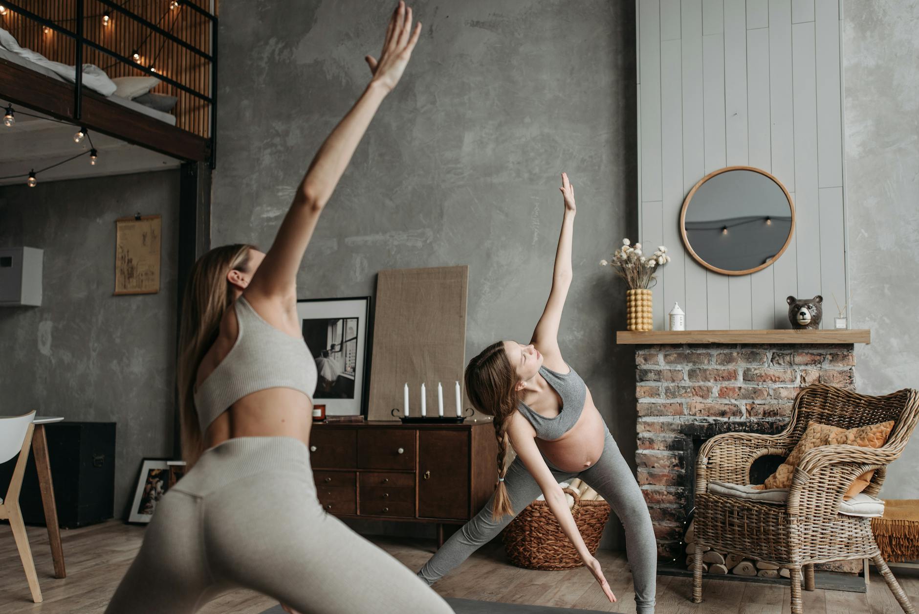 Two young women in a yoga studio. They are both wearing gray sports bras and leggings. The woman on the left is in a downward facing dog pose, with her arms stretched above her head and her legs bent at the knees. The other woman is standing on one leg, with the other leg extended behind her. In the background, there is a brick fireplace, a wicker chair, and a round mirror hanging on the wall. The room is decorated with string lights and various artworks. The overall aesthetic of the space is modern and minimalistic.
