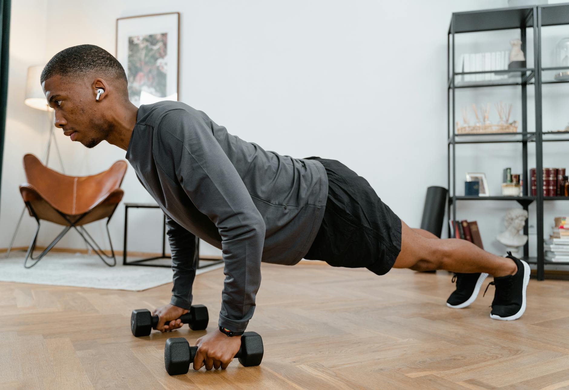 A young man performing a plank exercise with dumbbells in a living room. He is wearing a grey long-sleeved shirt and black sh...