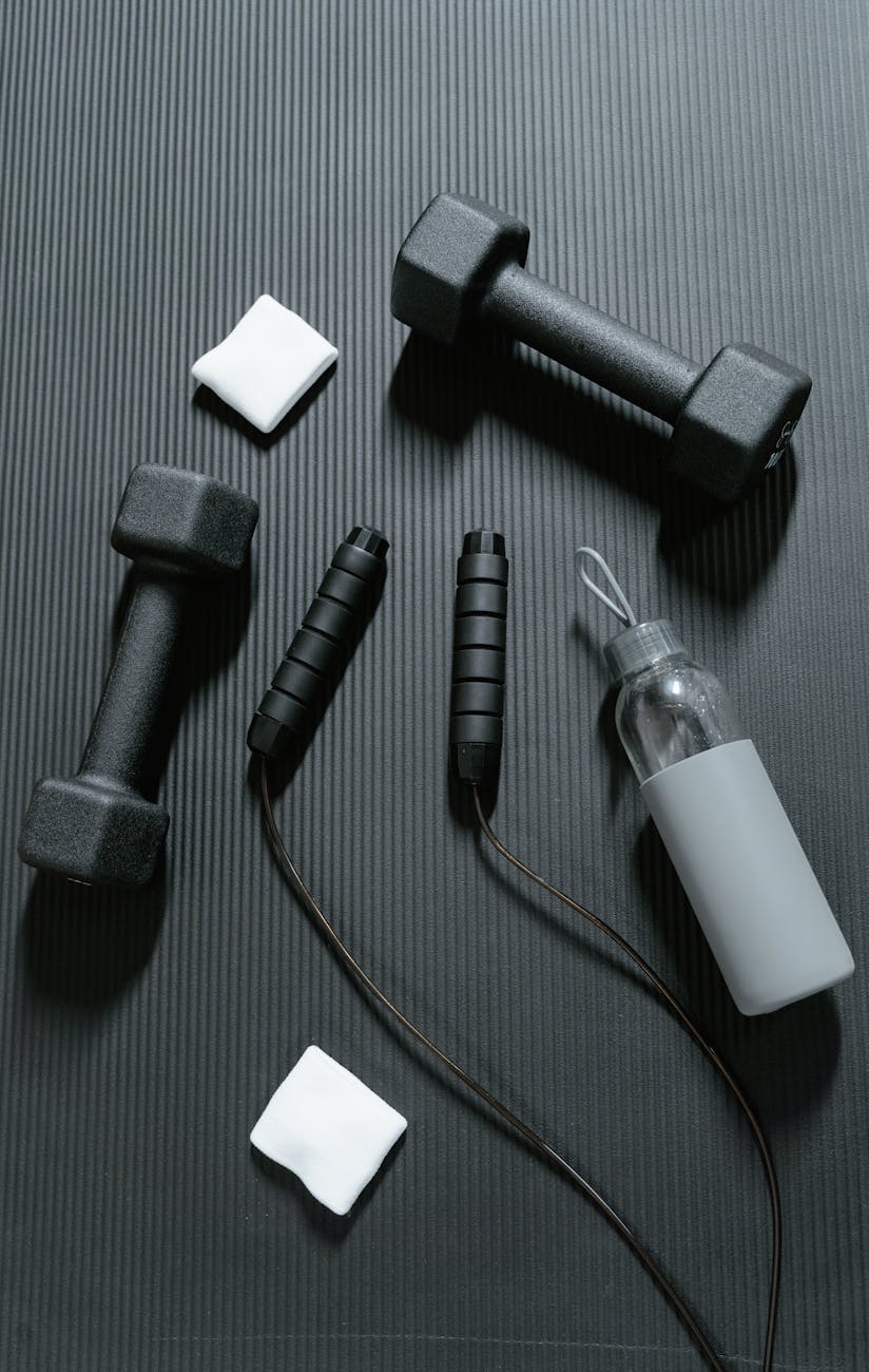 A set of fitness equipment laid out on a black textured surface. There are two black dumbbells on the left side of the image, one on the right side, and a small white square on the bottom left corner. Next to them, there is a black skipping rope with a black handle and a black cord attached to it. On the bottom right corner, there are two small white squares, one of which appears to be a water bottle. The items are arranged in a way that suggests they are being used for a workout or exercise routine.