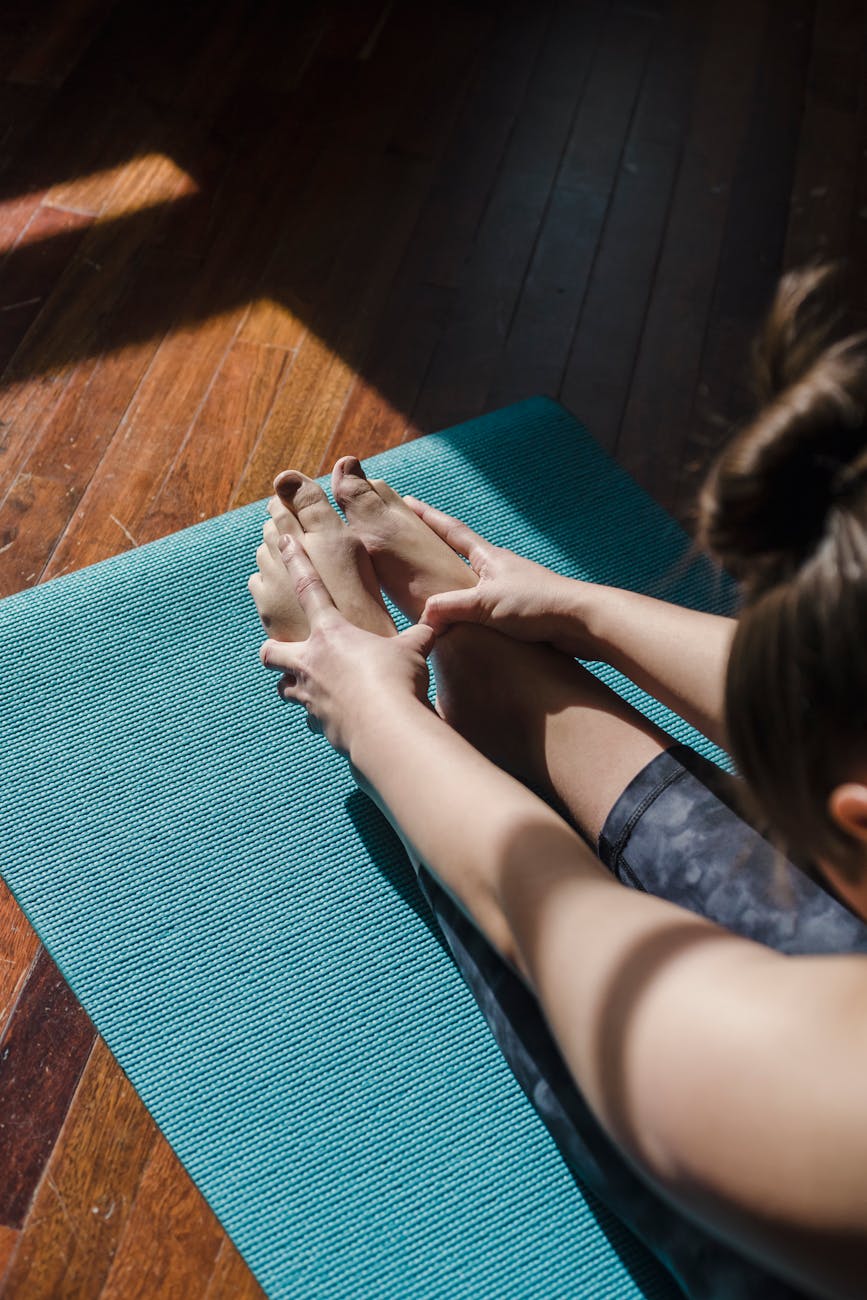 A person sitting on a blue yoga mat on a wooden floor. The person is holding their feet with their hands, and their legs are ...