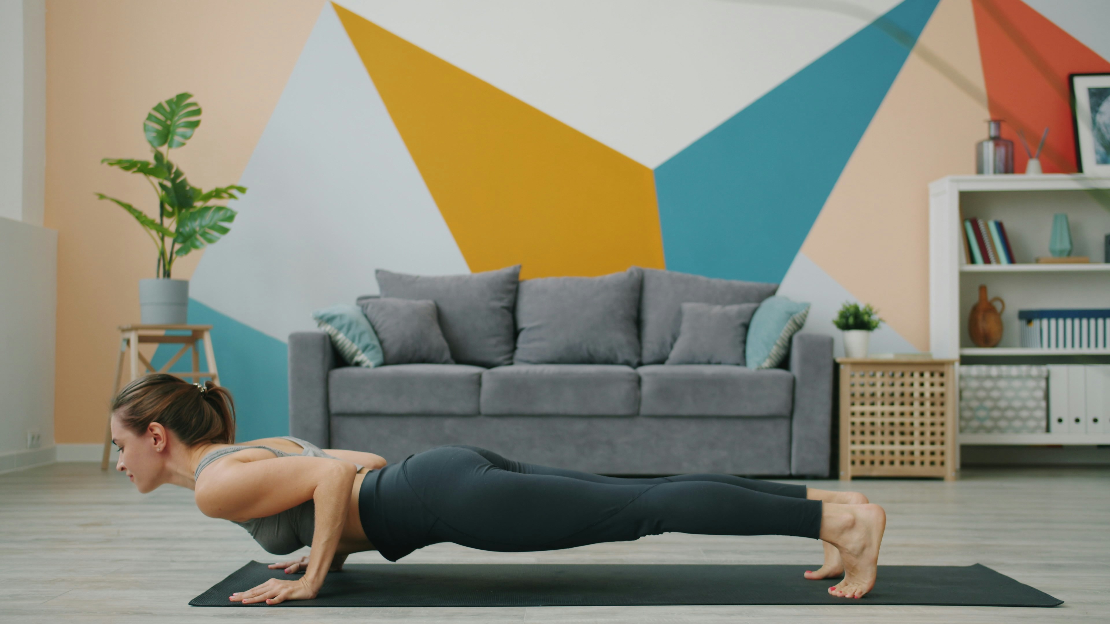 A young woman performing a plank exercise on a black yoga mat in a living room. She is wearing a gray tank top and black legg...