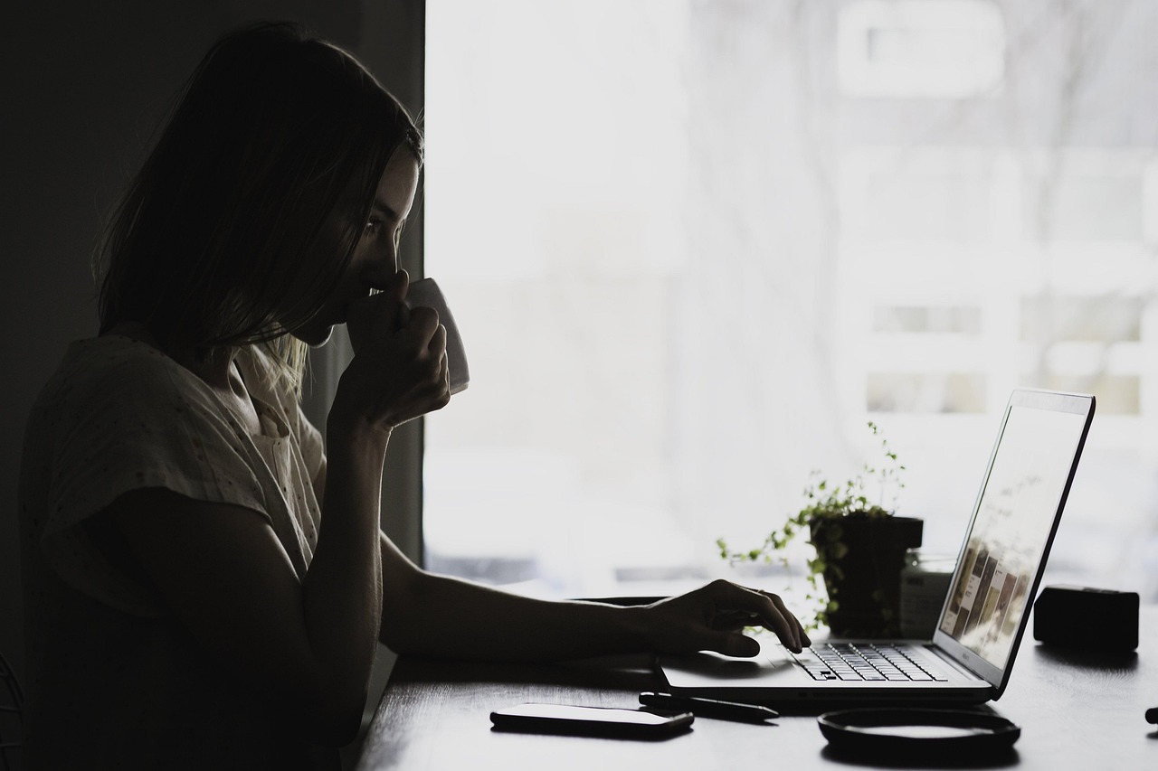 A young woman sitting at a desk with a laptop in front of her. She is holding a cup of coffee in her hand and appears to be d...