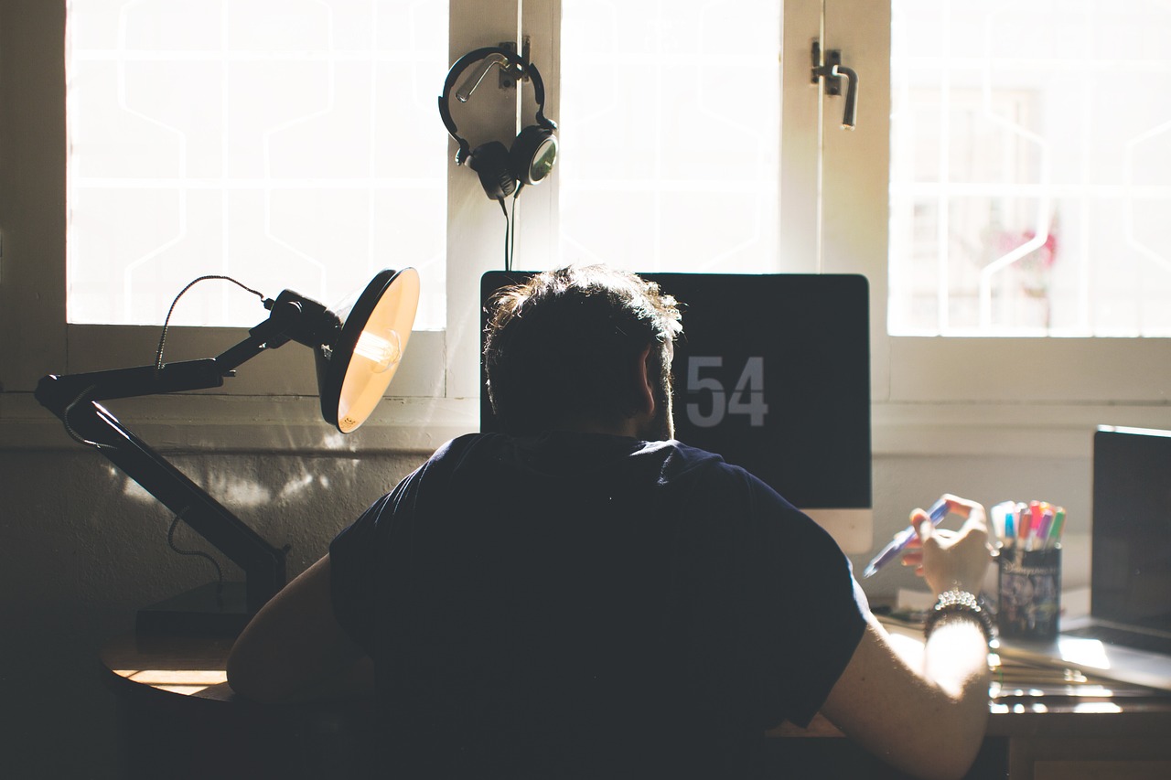 A man sitting at a desk in front of a computer monitor with the number 54 on it. He is wearing a black t-shirt and has a pair...