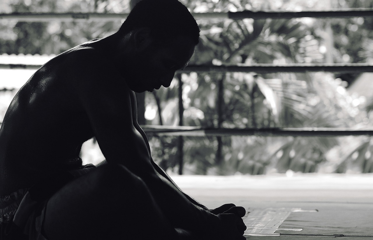 A black and white photograph of a man sitting on the floor in front of a window. He is shirtless and appears to be in a medit...