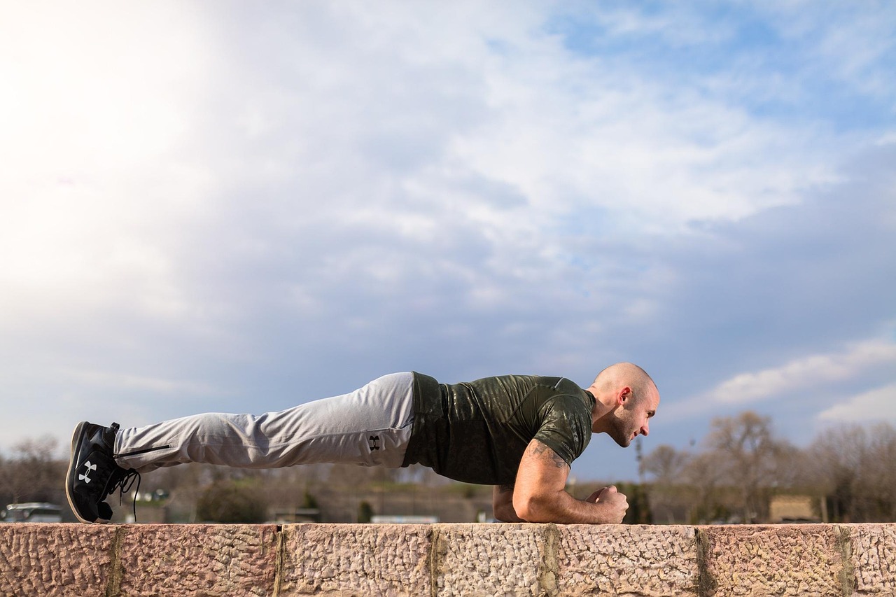 A man performing a plank exercise on a stone wall. He is wearing a green t-shirt, grey pants, and black sneakers. His arms ar...