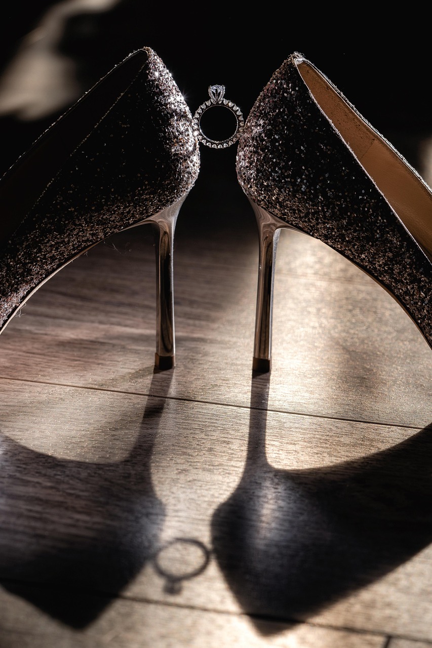 A pair of high-heeled shoes on a wooden floor. The shoes are black and silver in color and have a pointed toe and a stiletto heel. They are covered in a shiny, glittery material, giving them a luxurious and glamorous look. The background is blurred, making the shoes the focal point of the image. The focus is on the shoes, which are positioned in a way that they are casting a shadow on the floor, creating a sense of depth and dimension. The image is taken from a low angle, making them stand out against the dark background.