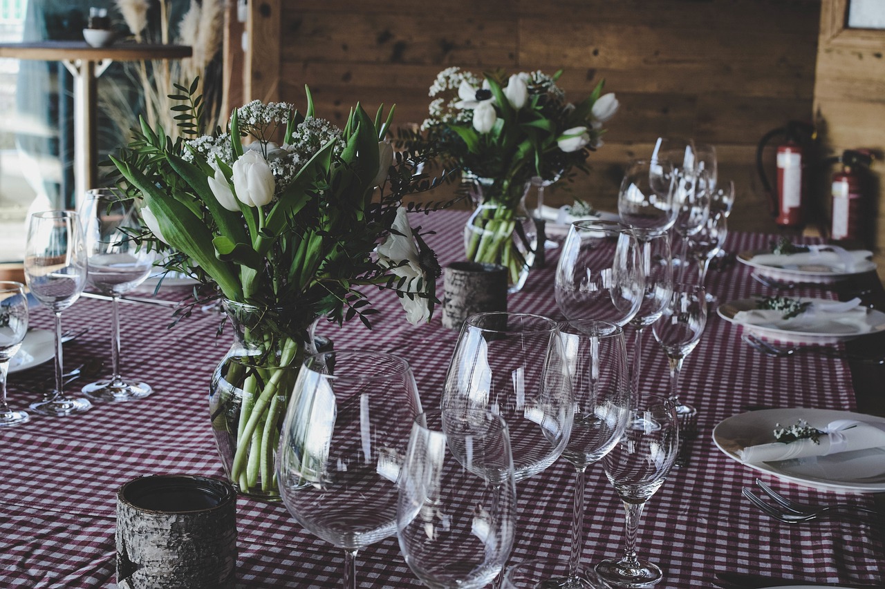 A long table set up for a formal dinner in a rustic setting. The table is covered with a red and white checkered tablecloth a...