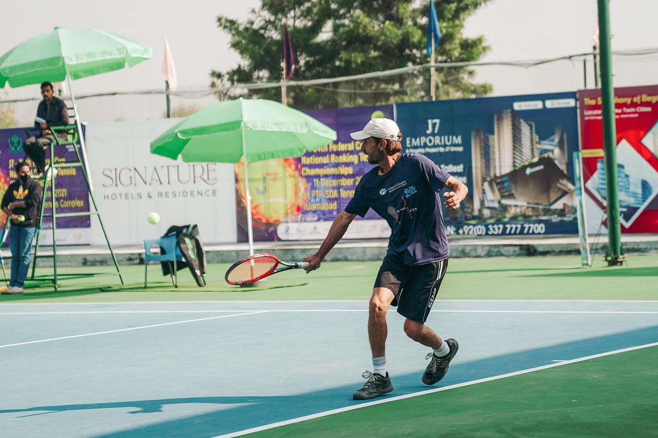A man playing tennis on a blue and green tennis court. He is wearing a blue t-shirt, black shorts, and a white cap. The man i...