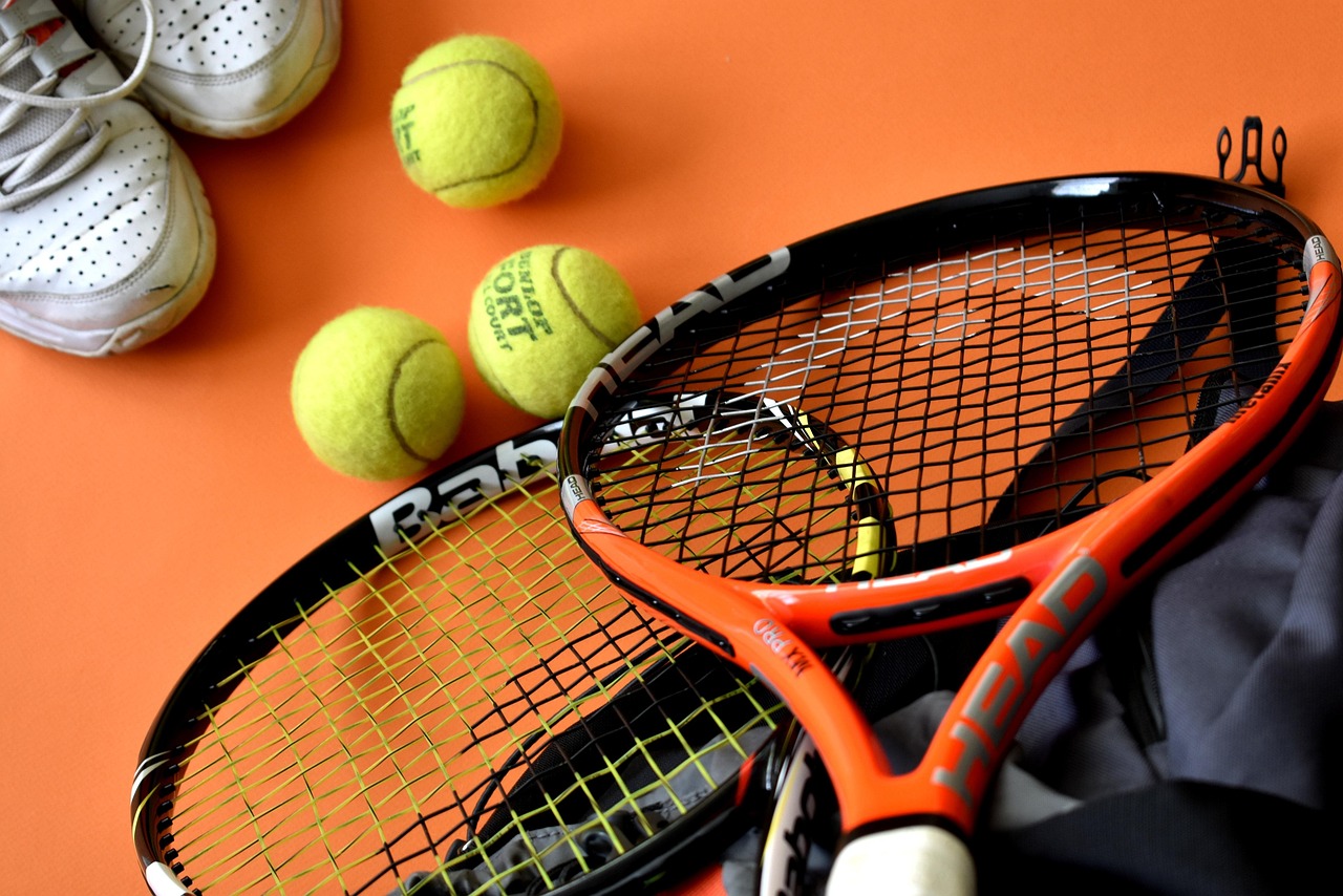 A close-up of a tennis racket and three tennis balls on an orange background. The racket is orange and black with the word "H...