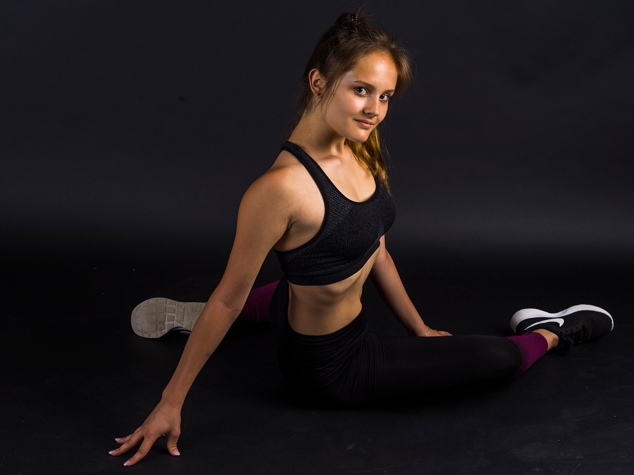 A portrait of a young woman sitting on the floor in a yoga pose. She is wearing a black sports bra and black leggings with pi...