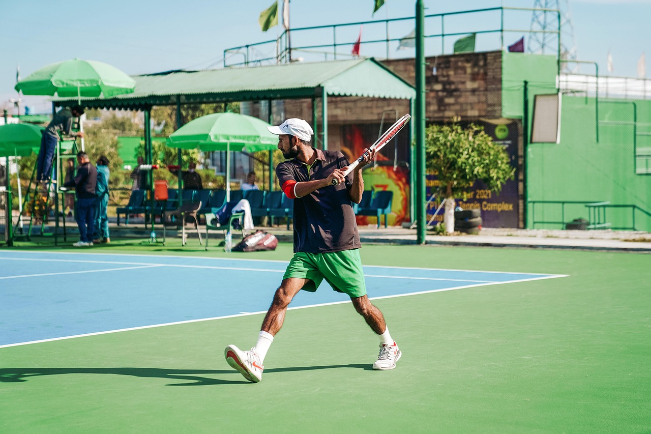 A man playing tennis on a blue and green tennis court. He is wearing a black t-shirt, green shorts, and white sneakers. He has a white cap on his head and is holding a red and white tennis racket in his hands. He appears to be in the middle of a swing, with his body slightly bent forward and his eyes focused on the ball. In the background, there are green umbrellas and a few people watching the game. The sky is blue and there are a few clouds in the sky.