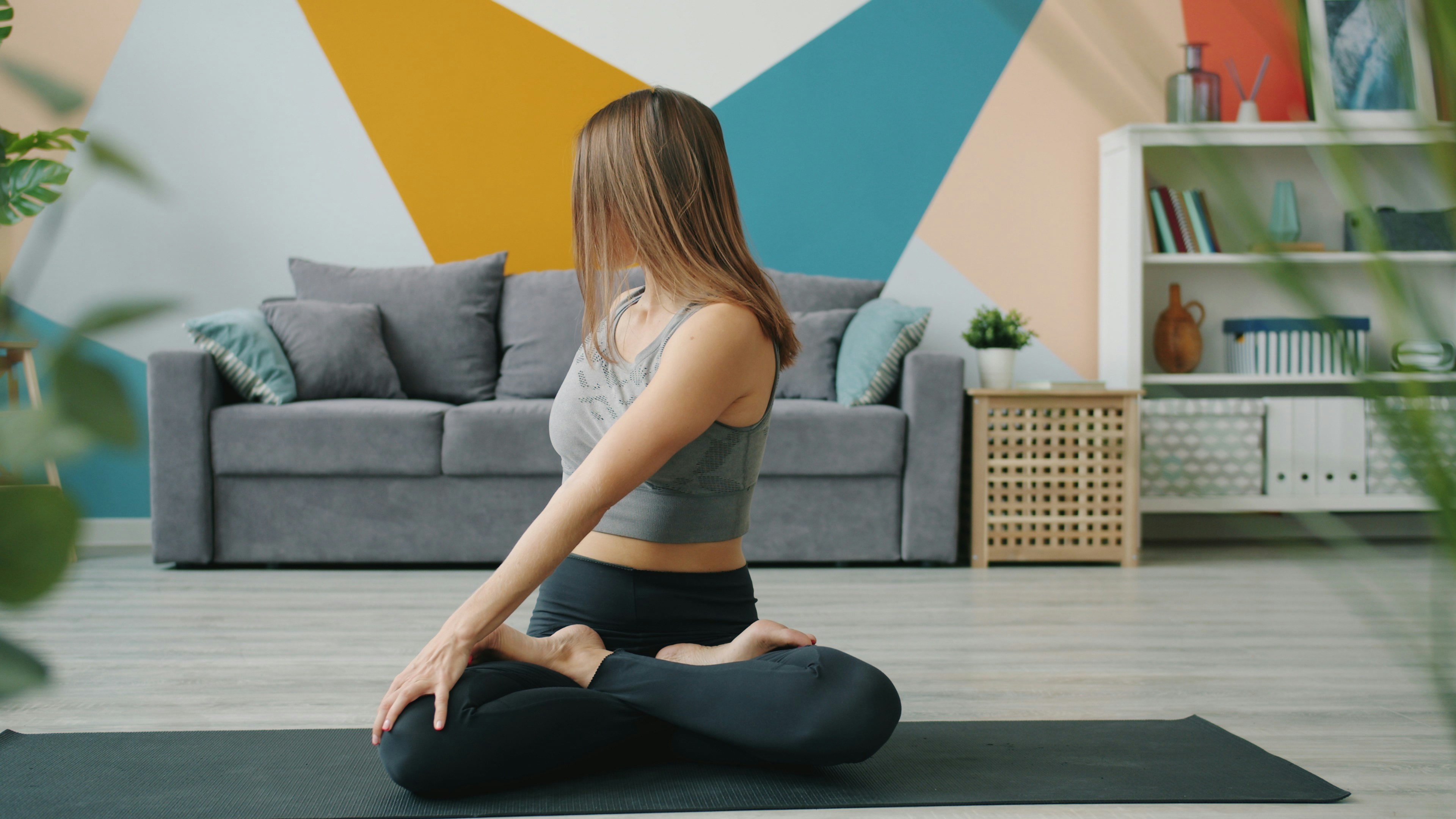 A young woman sitting on a black yoga mat in a living room. She is wearing a gray sports bra and black leggings and is in a m...