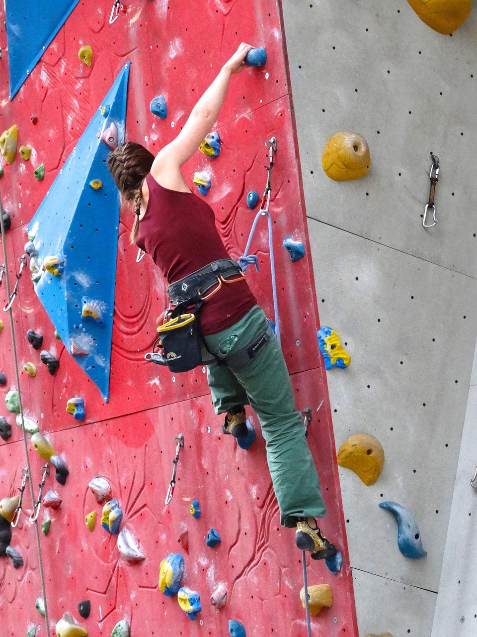 A young woman rock climbing on an indoor rock wall. She is wearing a maroon tank top, green pants, and black shoes. She has a...