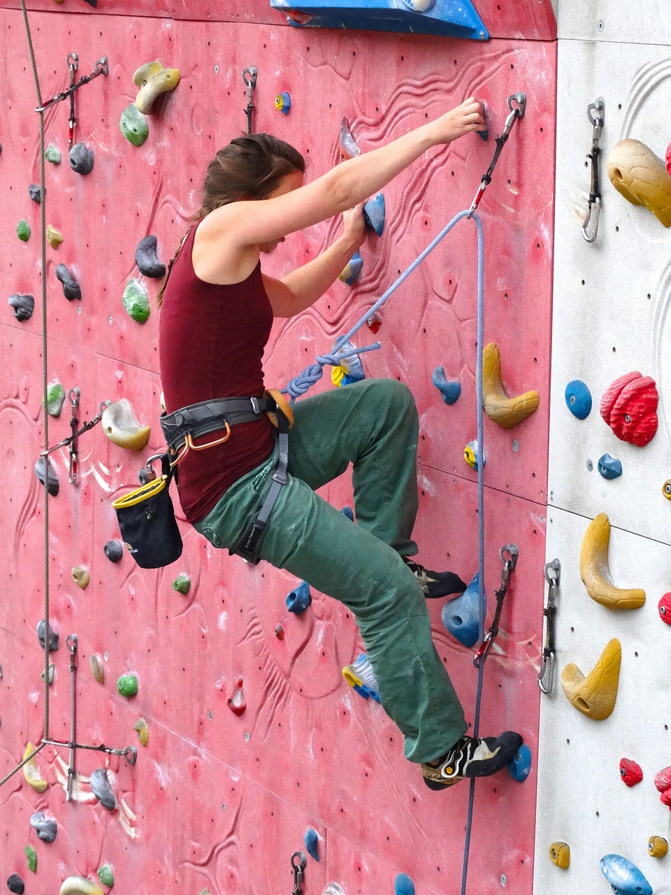 A young woman rock climbing on an indoor rock wall. She is wearing a maroon tank top, green pants, and black shoes. She has a...