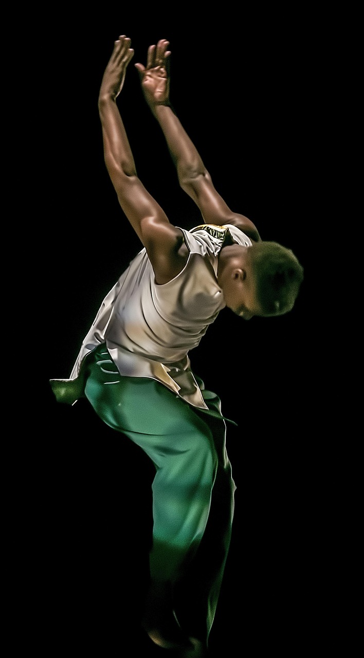 A young man in mid-air, performing a dance move. He is wearing a white tank top and green pants. His arms are stretched upwar...