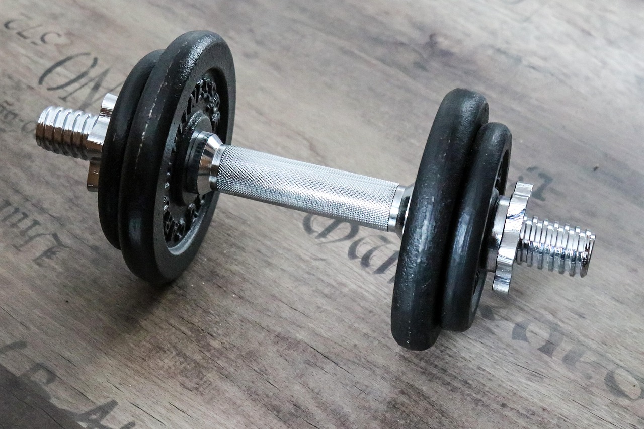 A pair of dumbbells lying on a wooden surface. The dumbbell on the left is black and appears to be made of rubber, while the one on the right is silver. Both weights have a hexagonal shape with a threaded end and a flat base. The handle of the dumbbell is made of metal and has a textured surface for better grip. The background is blurred, but it seems to be a wooden table or countertop.