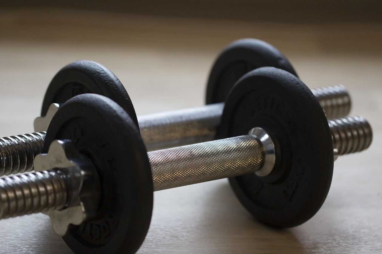 A pair of dumbbells lying on a wooden surface. The dumbbell on the left is black and appears to be made of rubber, while the ...
