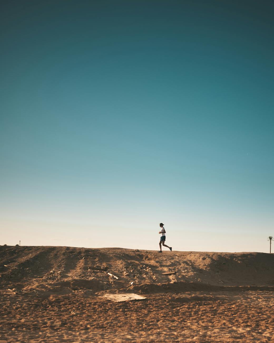 A person walking on a sandy hill with a clear blue sky in the background. The person is wearing a white shirt and black pants...
