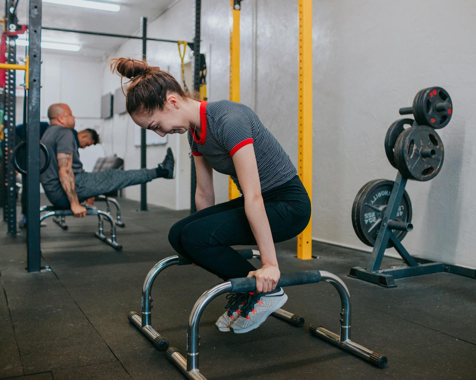 A young woman in a gym performing a bench press exercise. She is wearing a black and red striped shirt, black leggings, and w...