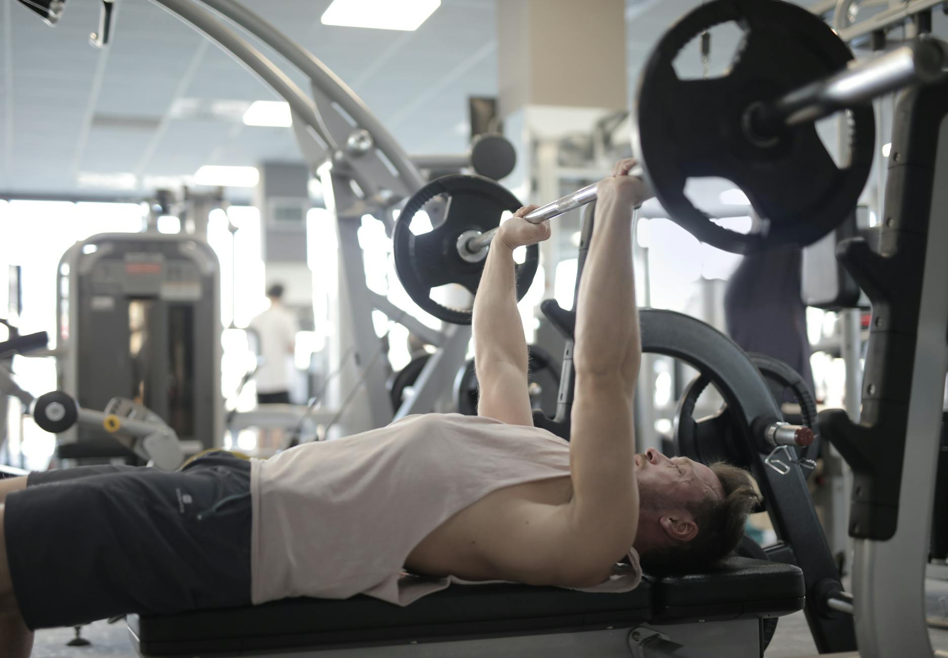 A man performing a bench press exercise in a gym. He is lying on a black bench with his feet shoulder-width apart and his arm...