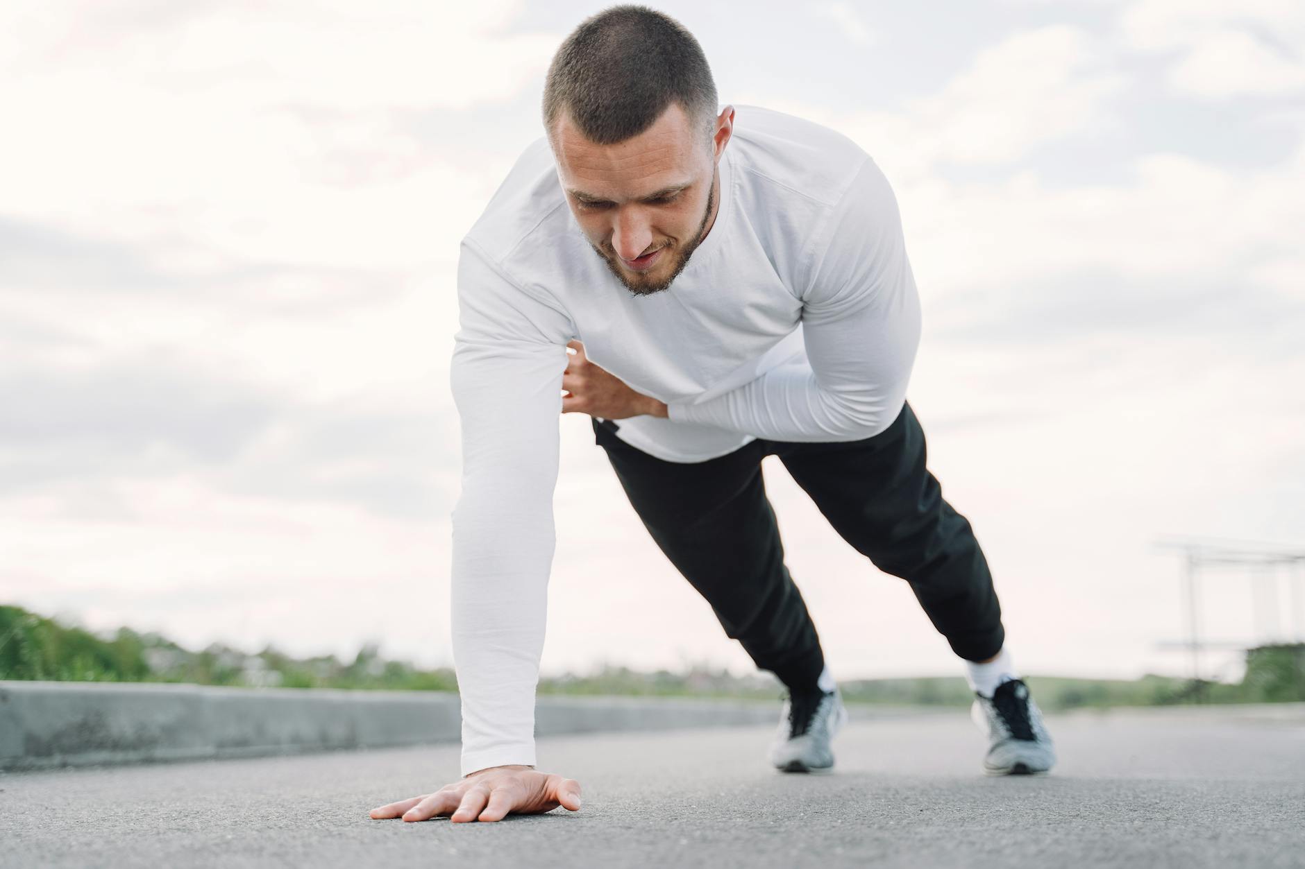 A young man in a white long-sleeved shirt and black pants doing push-ups on an empty road. He is in a crouched position with ...