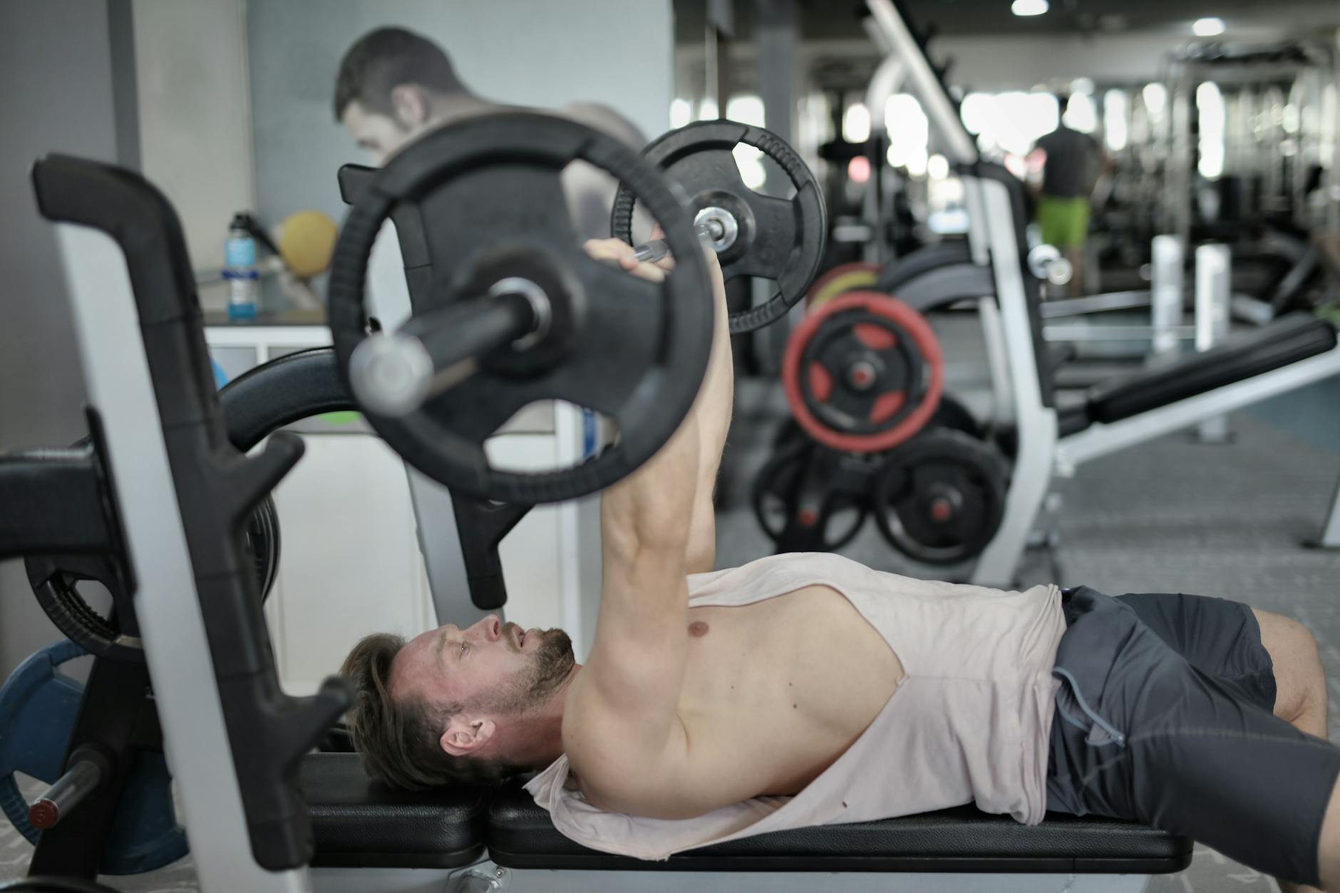 A man lying on a bench in a gym, performing a bench press exercise. He is holding a barbell above his head with both hands an...