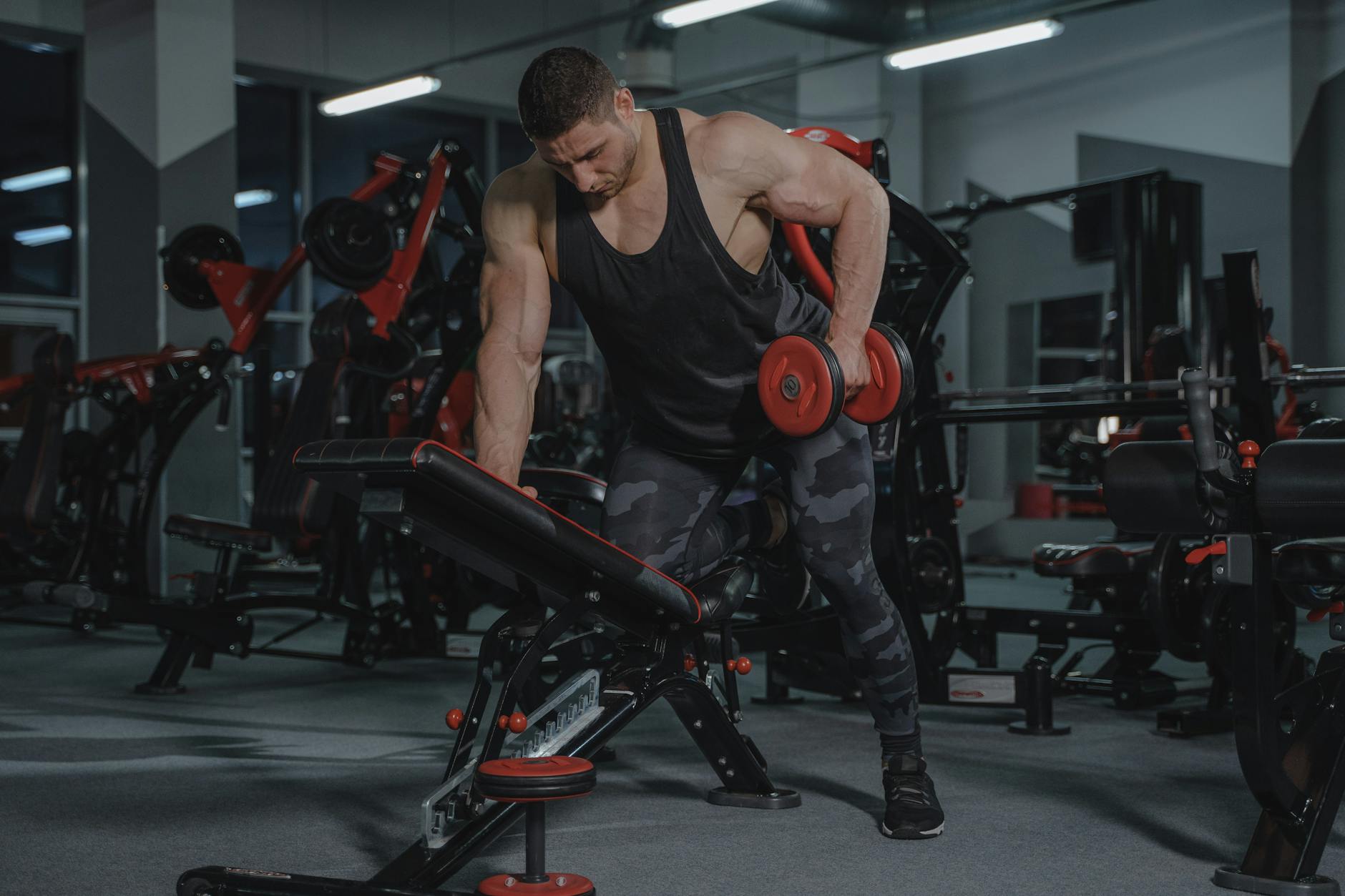 A young man working out in a gym. He is wearing a black tank top and camouflage pants and is using a bench press machine with...
