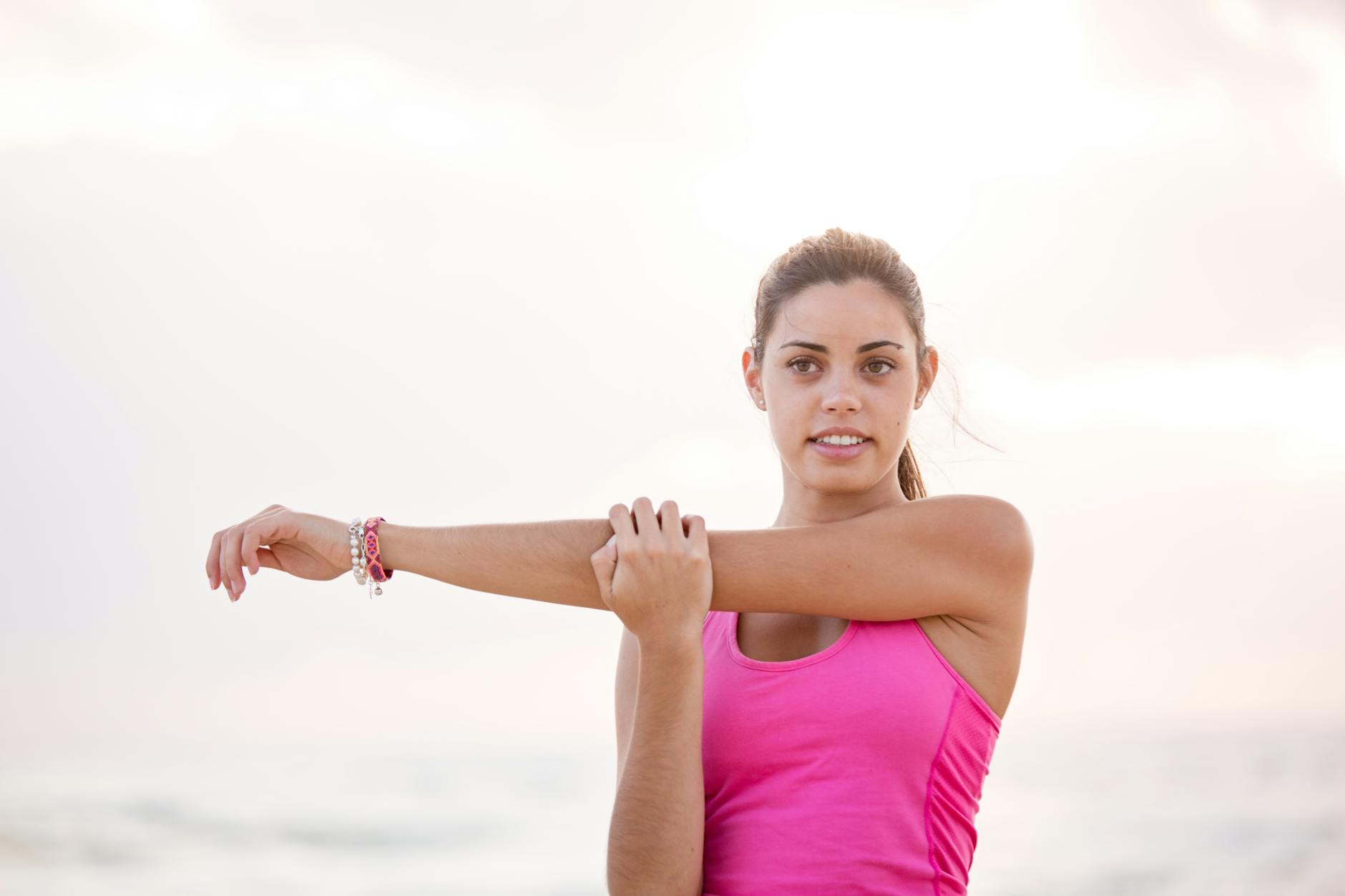 A young woman standing on a beach with the ocean in the background. She is wearing a pink tank top and has her arms stretched...
