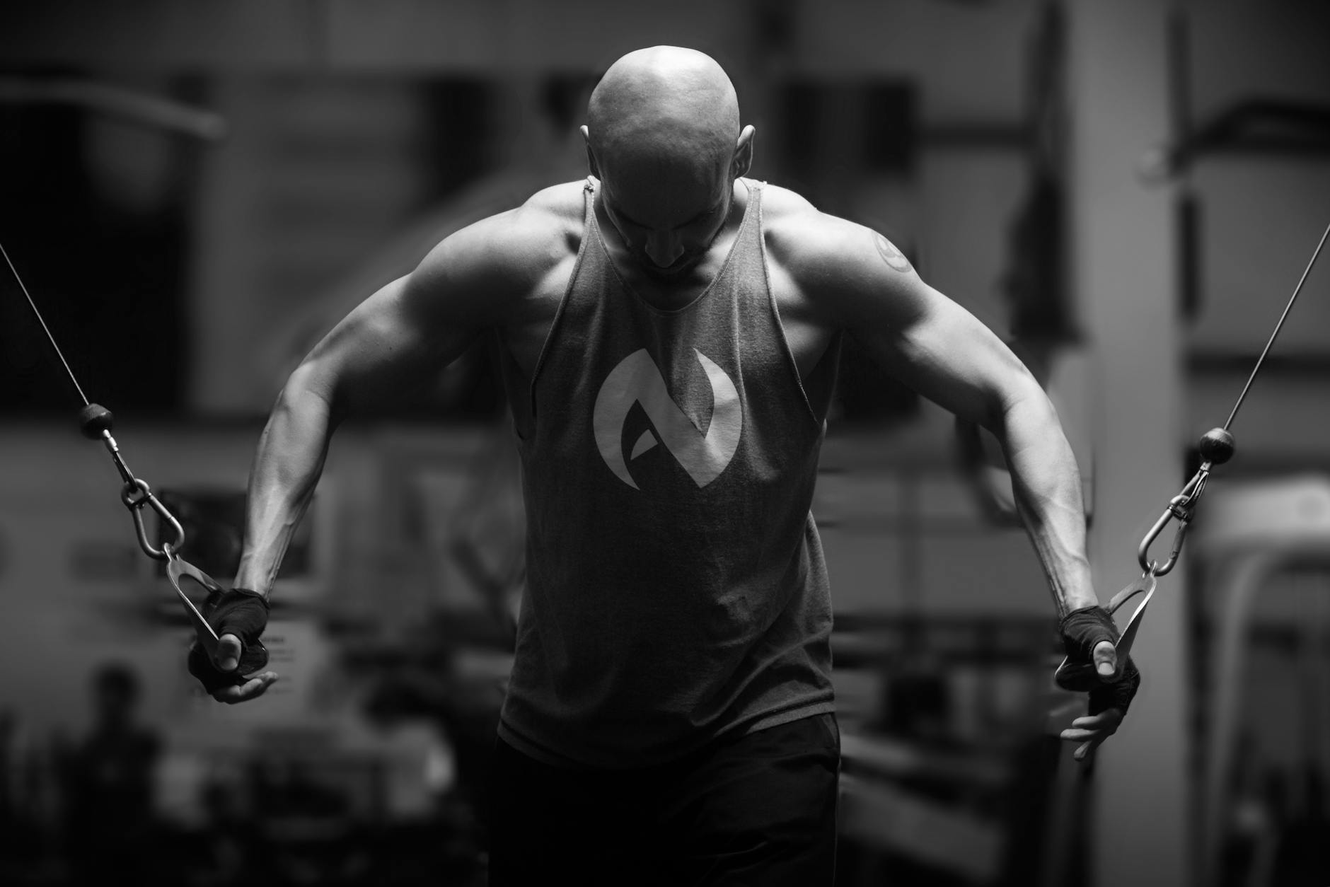 A black and white photograph of a man working out in a gym. He is wearing a sleeveless tank top with a white logo on it and b...