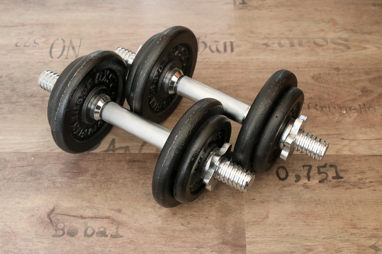A pair of dumbbells lying on a wooden surface. The dumbbell on the left is black and appears to be made of rubber, while the ...