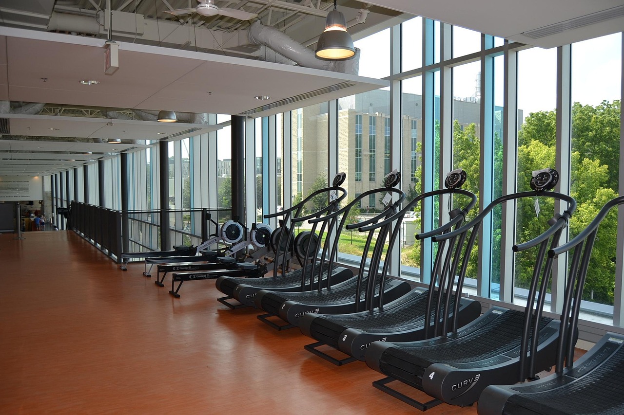 A row of treadmills in a modern gym. The gym has a high ceiling with large windows that offer a view of the city skyline. The...