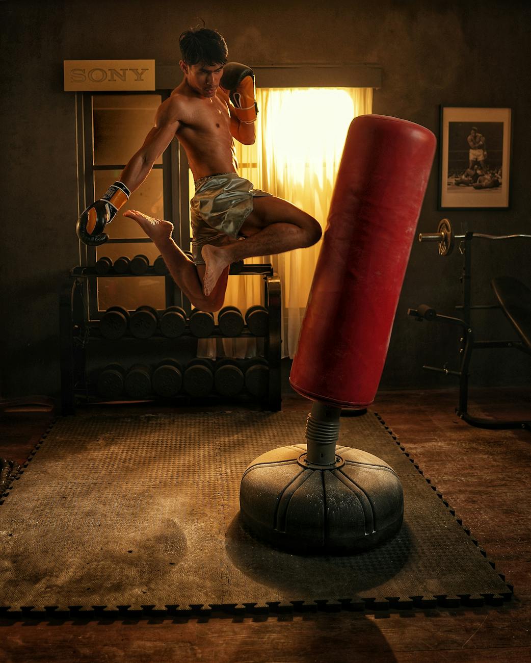 A young man in a gym, performing a kickboxing exercise. He is wearing shorts and boxing gloves and is in mid-air, with his le...