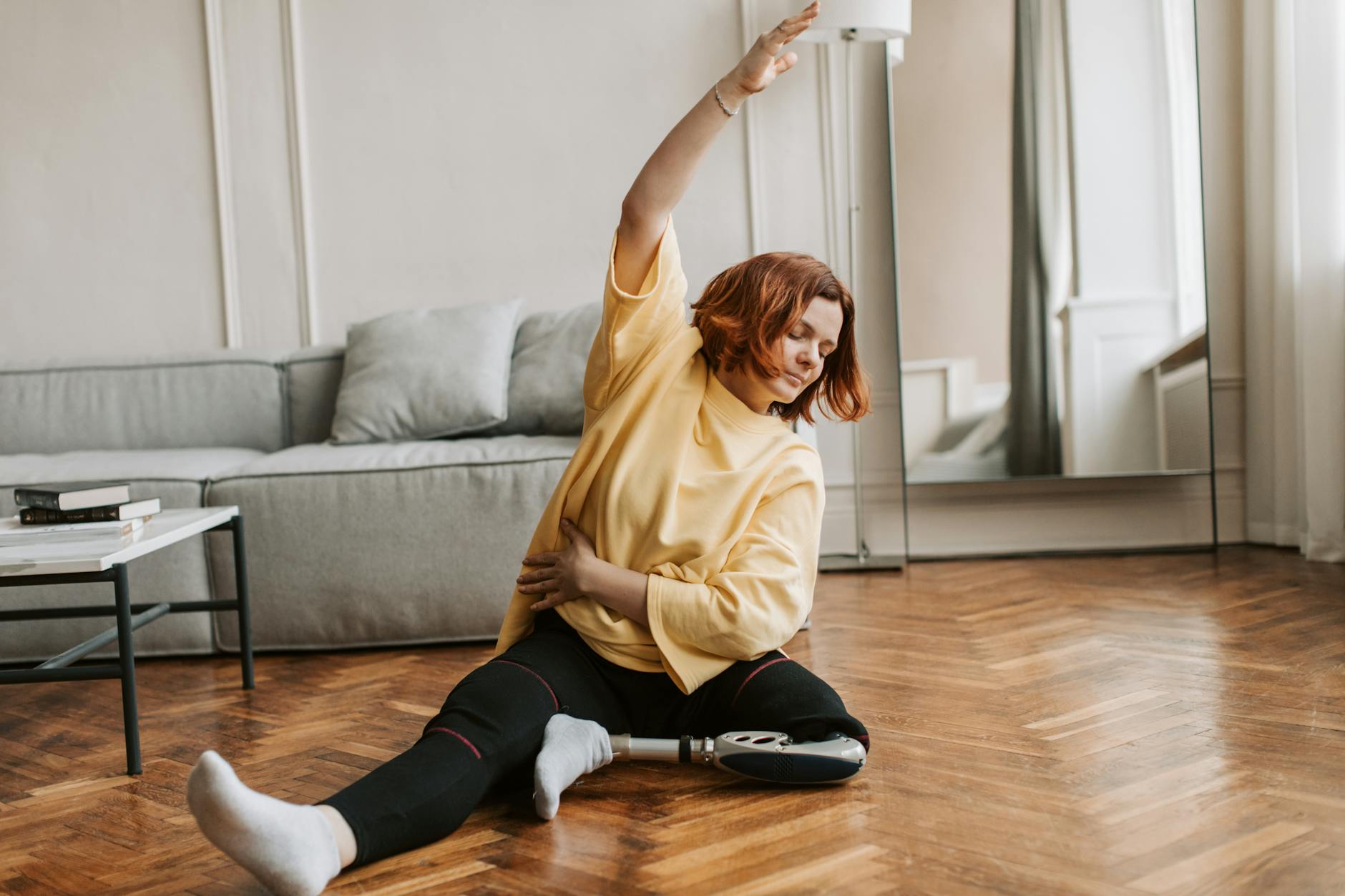 A young woman sitting on the floor in a living room. She is wearing a yellow sweatshirt, black leggings, and white socks. Her...