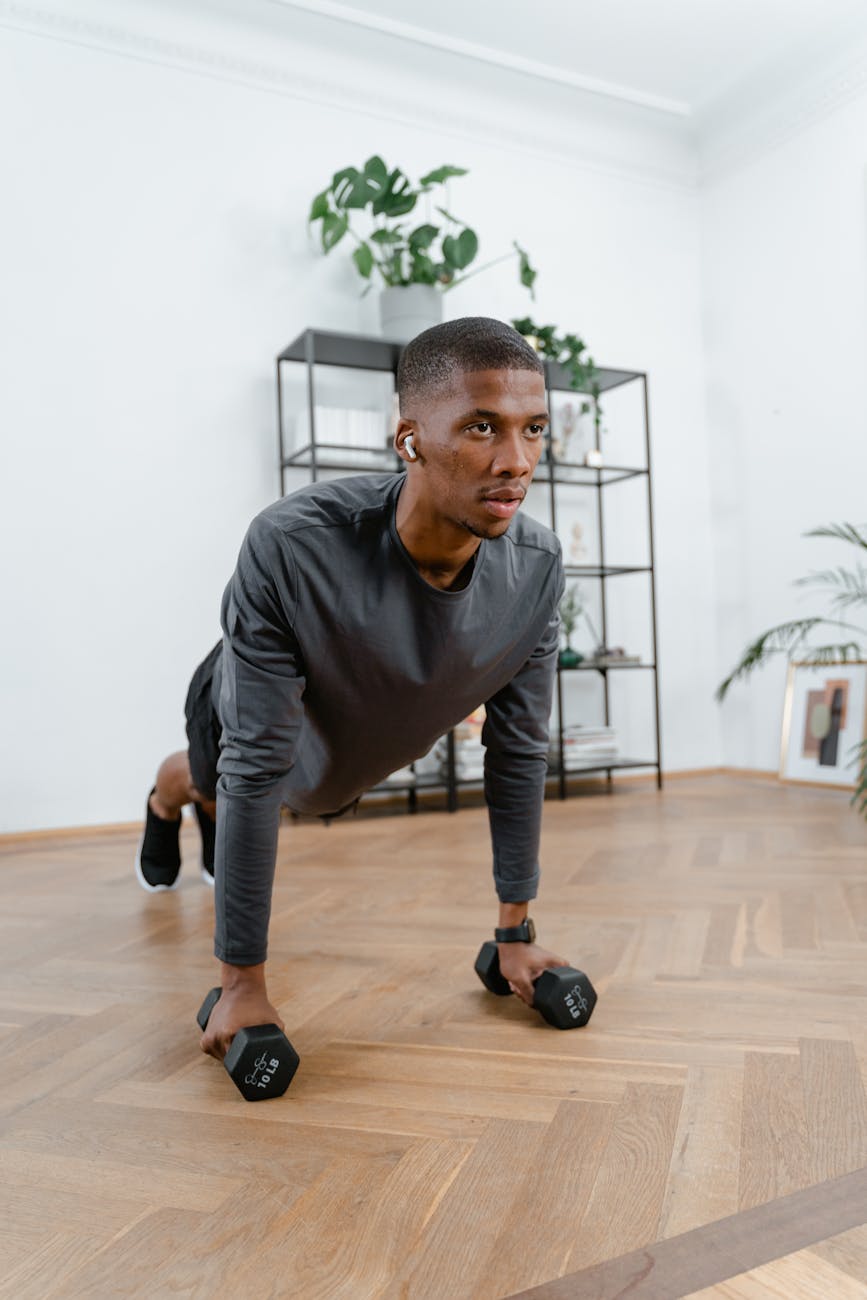A young man in a gym, performing a push-up exercise with dumbbells. He is wearing a grey long-sleeved shirt and black shorts,...