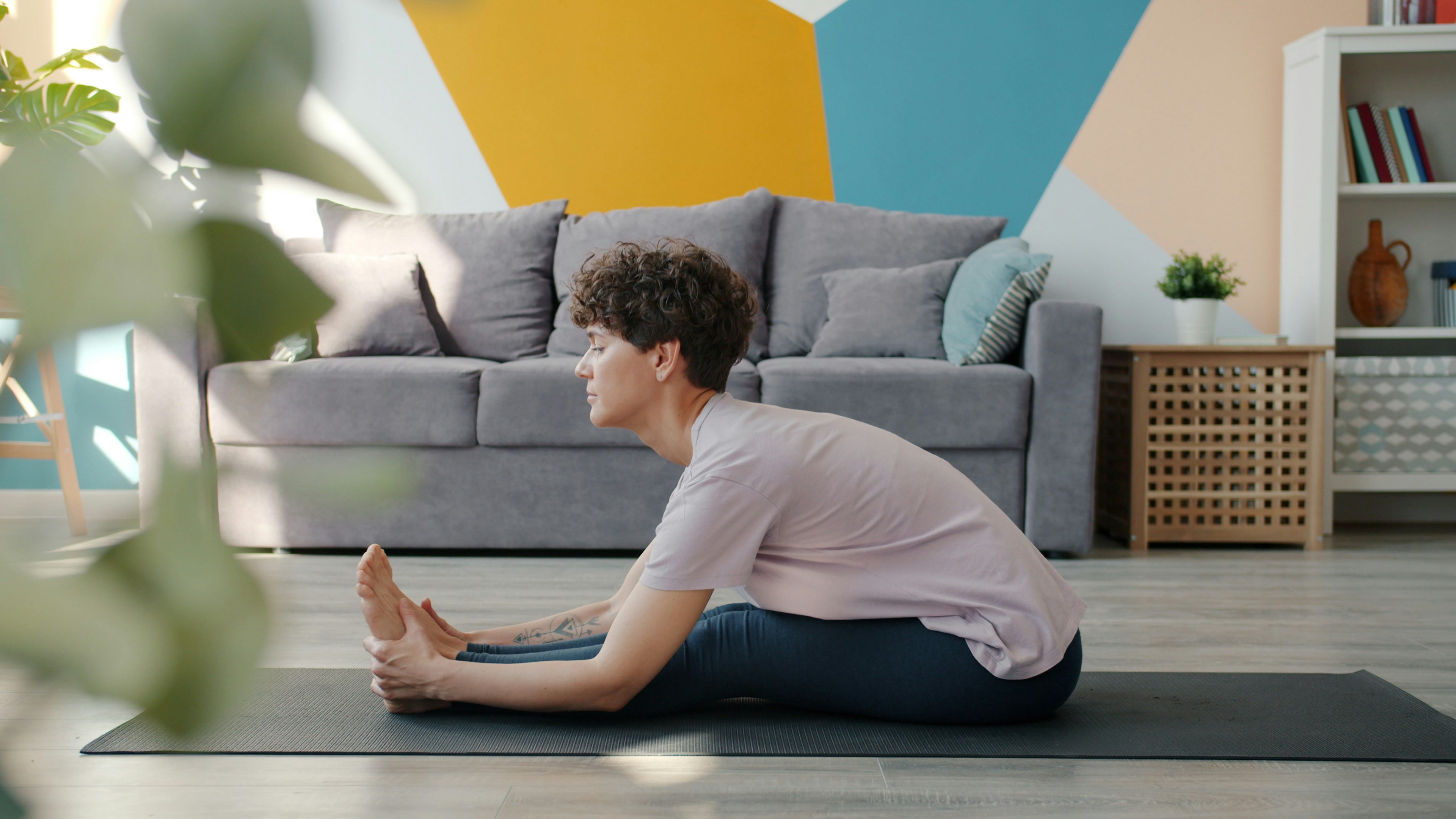 A young woman sitting on a black yoga mat in a living room. She is wearing a pink t-shirt and blue leggings and has short cur...