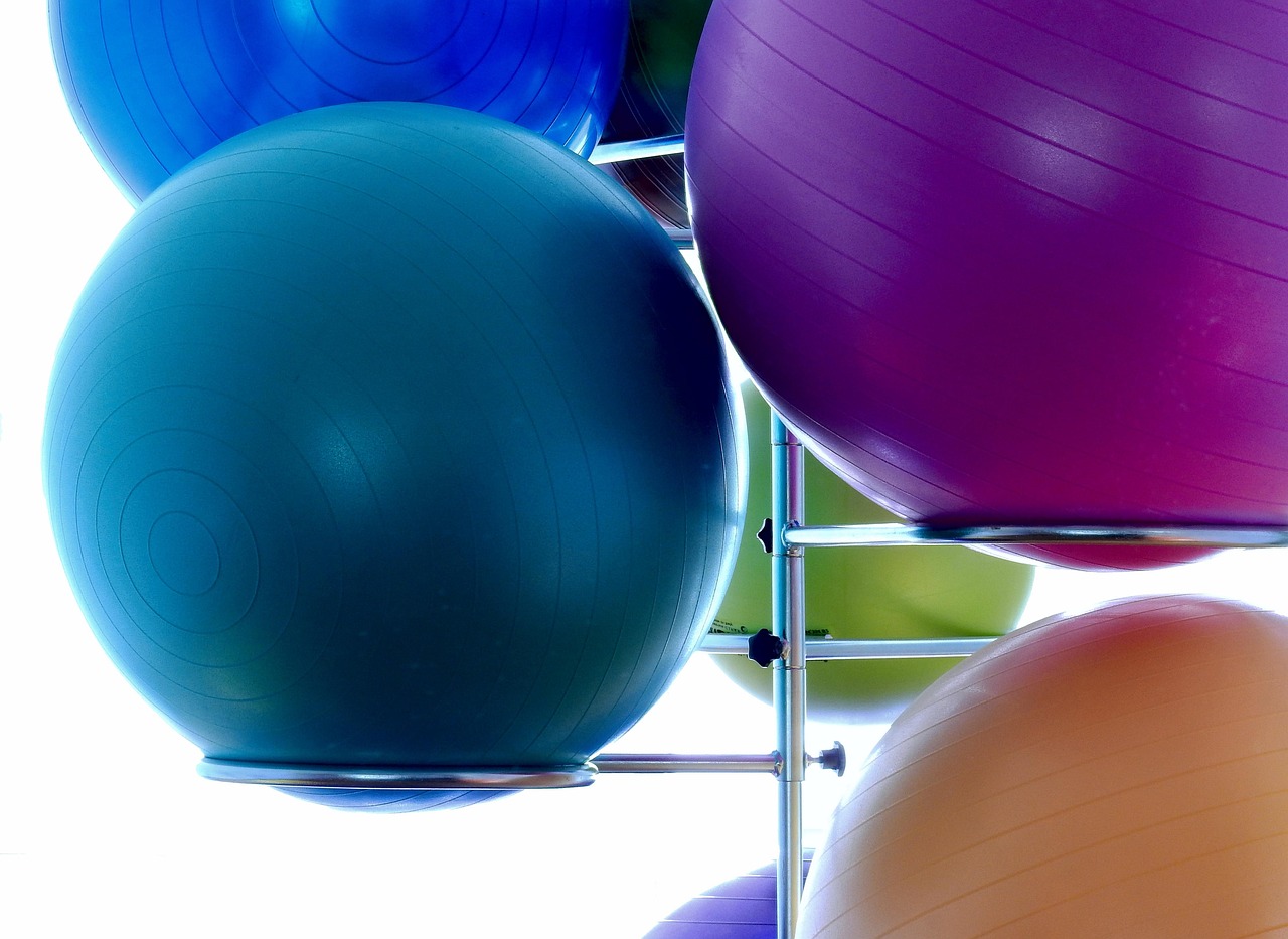 A group of exercise balls of different colors - blue, purple, green, orange, and yellow - hanging from a metal rack. The balls are arranged in a way that they are overlapping each other, creating a sense of depth and dimension. The background is white, making the colors of the balls stand out even more. The image appears to be taken from a low angle, looking up at the balls.