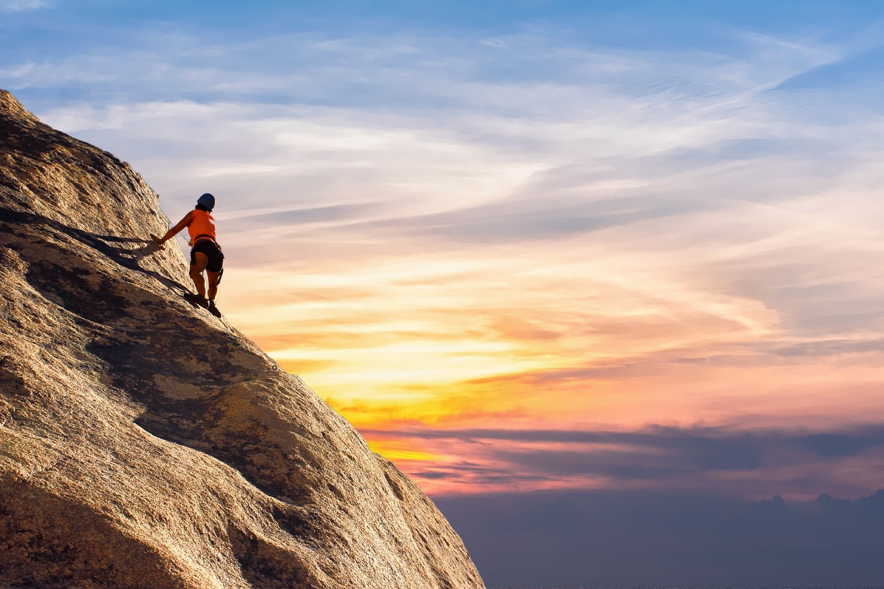 A person rock climbing on a cliff at sunset. The person is wearing an orange shirt and black shorts and is holding onto a rope for support. The cliff is made of large rocks and the sky is a beautiful orange and pink color, with the sun setting in the background. The sky is filled with clouds and the horizon is visible in the distance. The overall mood of the image is peaceful and serene.