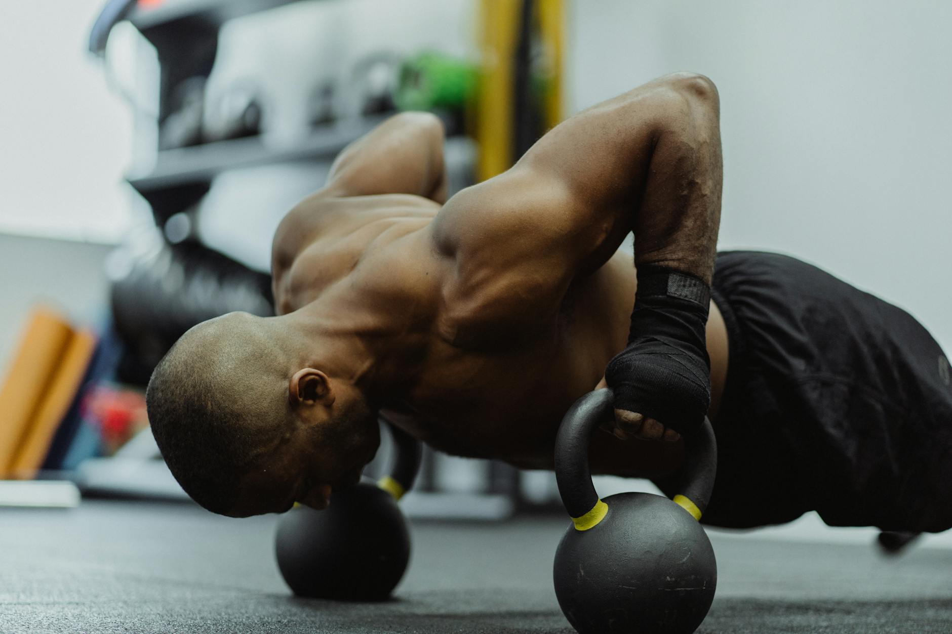 A man performing a kettlebell push-up exercise in a gym. He is lying on his back with his feet shoulder-width apart and his a...