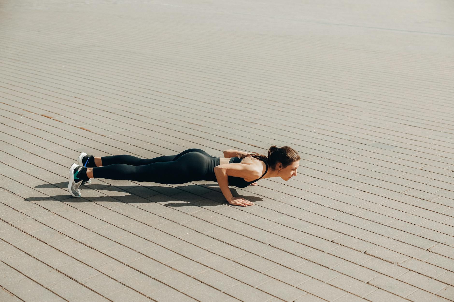 A young woman performing a plank exercise on a brick pavement. She is wearing a black sports bra and black leggings, and whit...