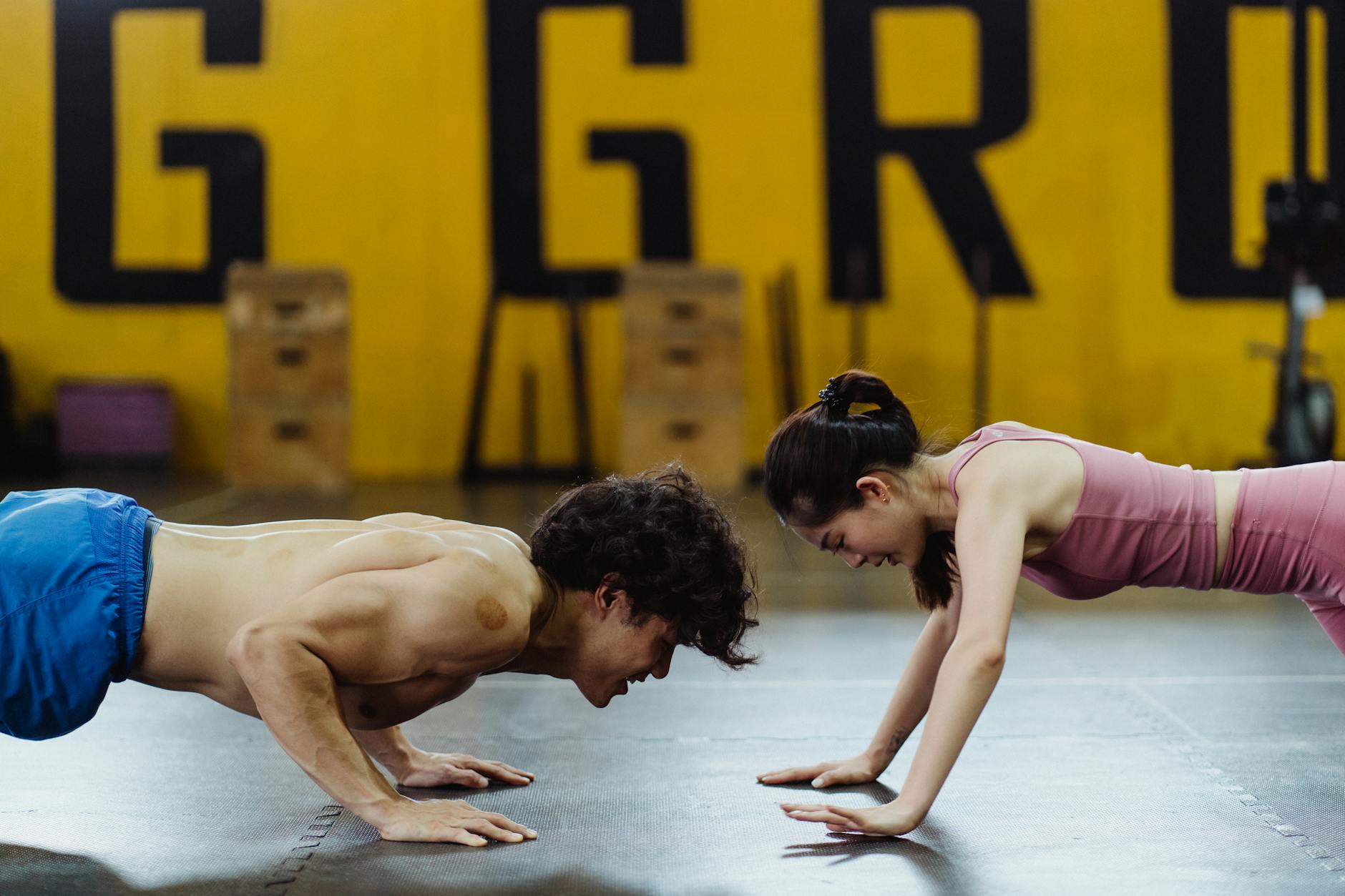 A man and a woman in a gym, performing a plank exercise. The man is shirtless and wearing blue shorts, while the woman is wea...