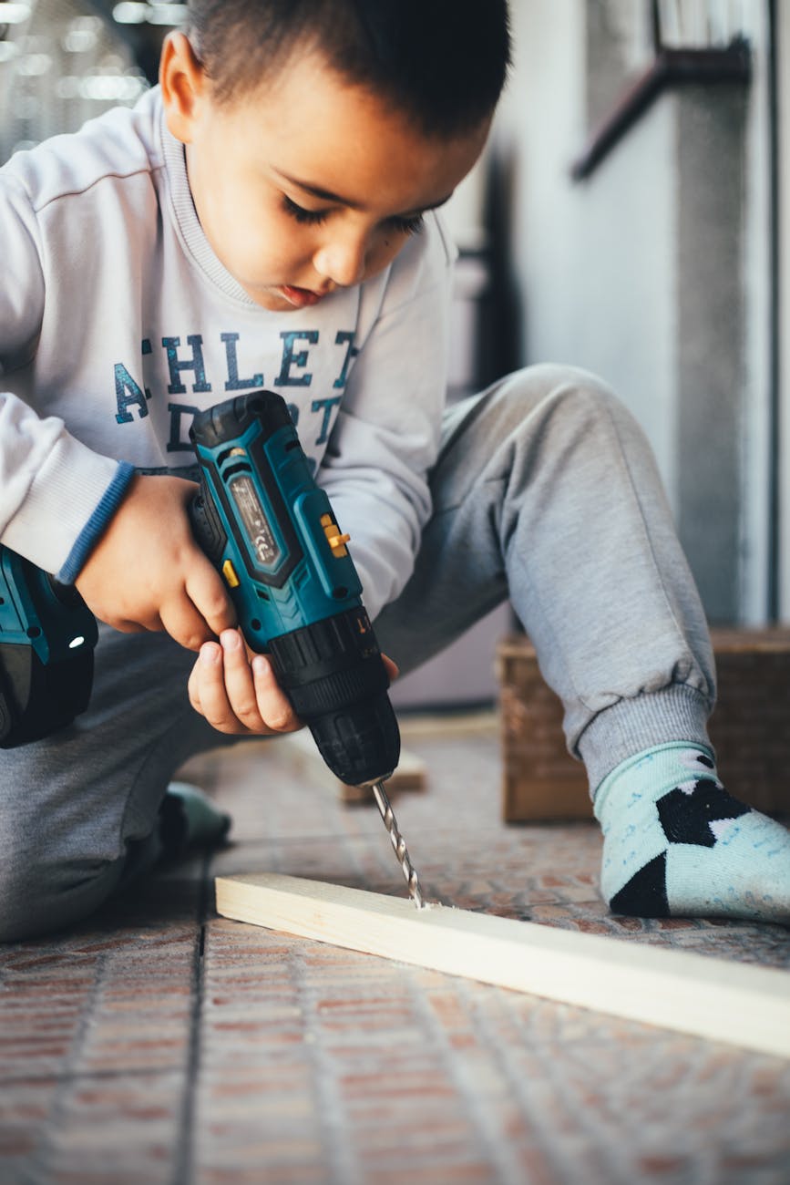 A young boy, probably around 4-5 years old, sitting on the floor and using a power drill to drill a piece of wood. He is wear...