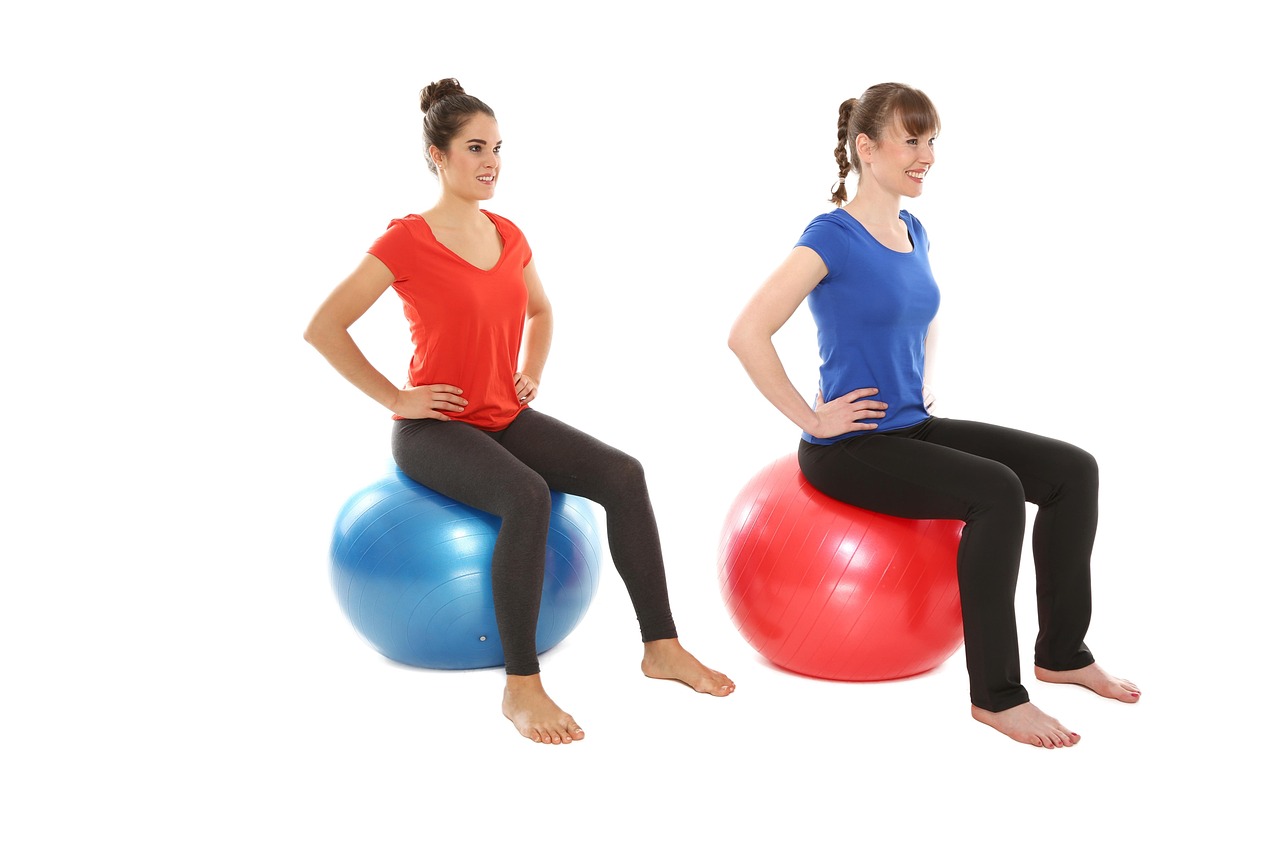 Two young women sitting on exercise balls. The woman on the left is wearing a red t-shirt and black leggings. She is sitting ...